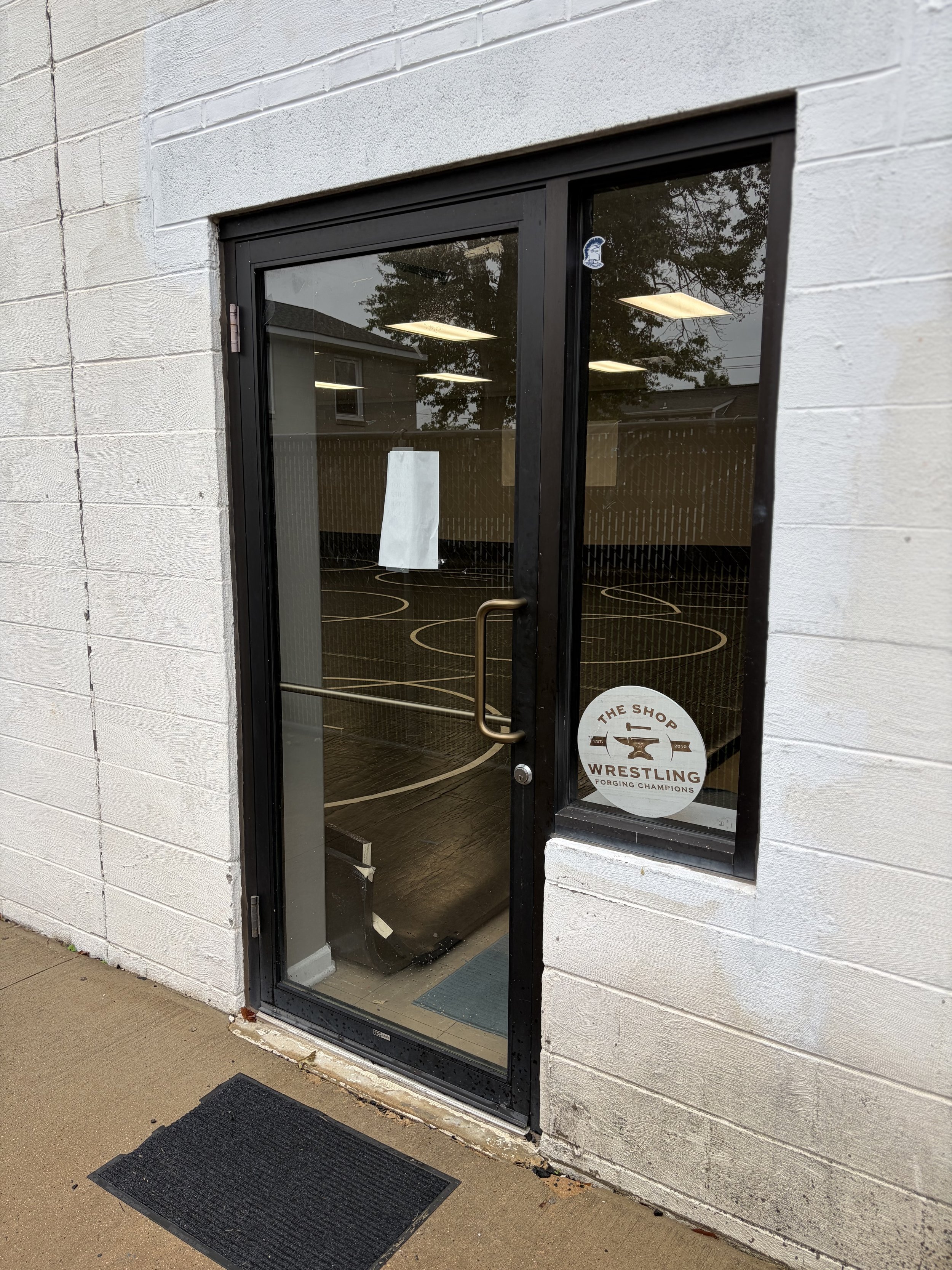 Exterior view of a glass door with a round sign that reads 'The Shop Wrestling Forging Champions,' and the reflection of trees and buildings in the glass. The door is set in a white brick wall with a black metal frame.