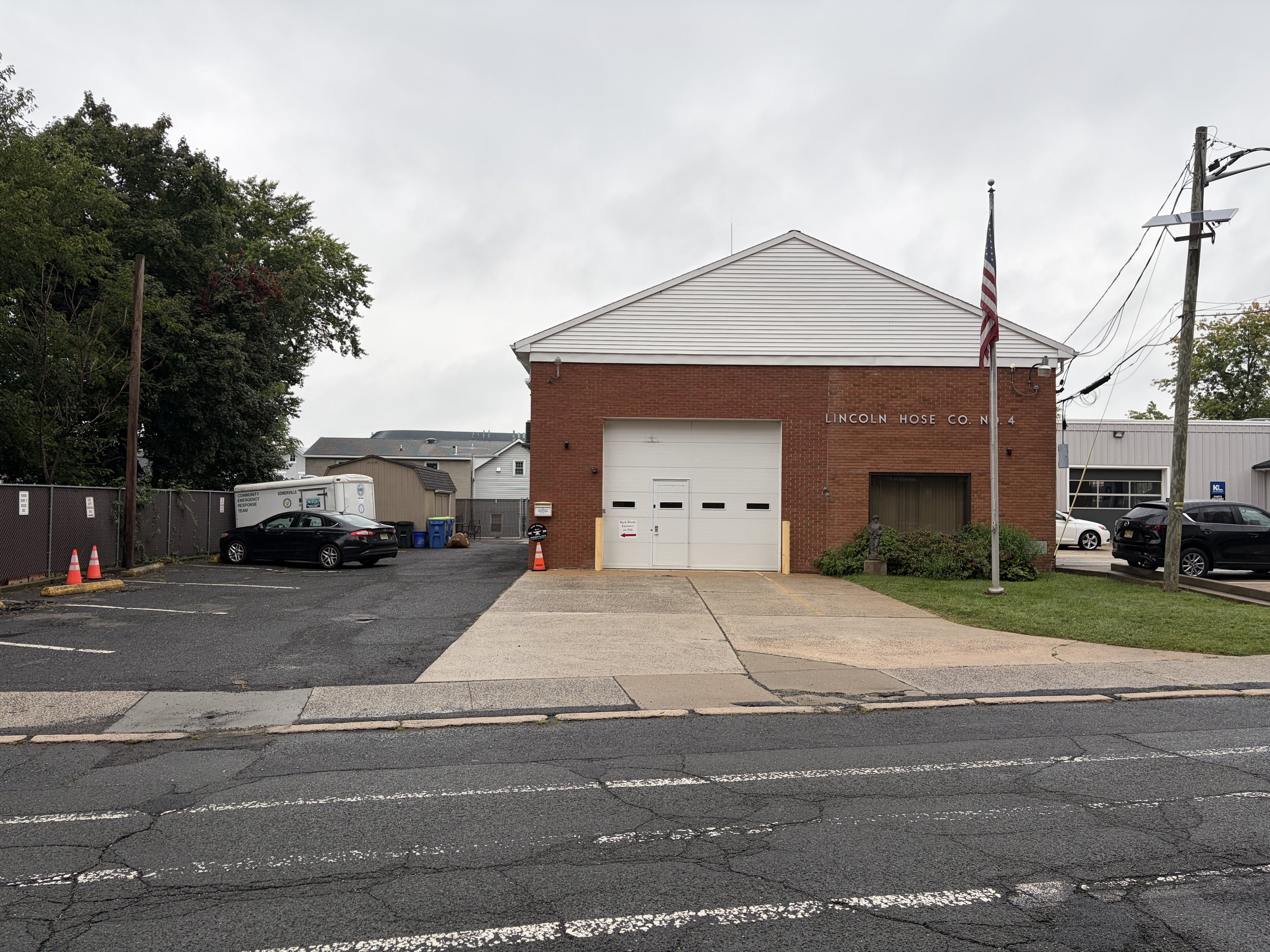 View of a building with a sign that reads 'Lincoln Hose Co. N 4,' a gray sky, trees, parked cars, a utility pole with wires and a flag, a fenced area, and a parking lot with some traffic cones.