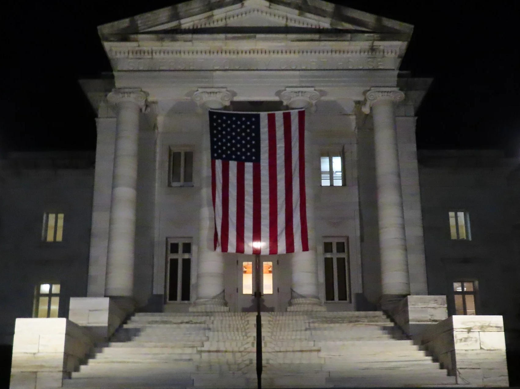 Nighttime view of a government building with Greek Revival architecture, featuring tall columns and a pediment; an American flag is hung vertically in front of the entrance, and the building is illuminated from within.
