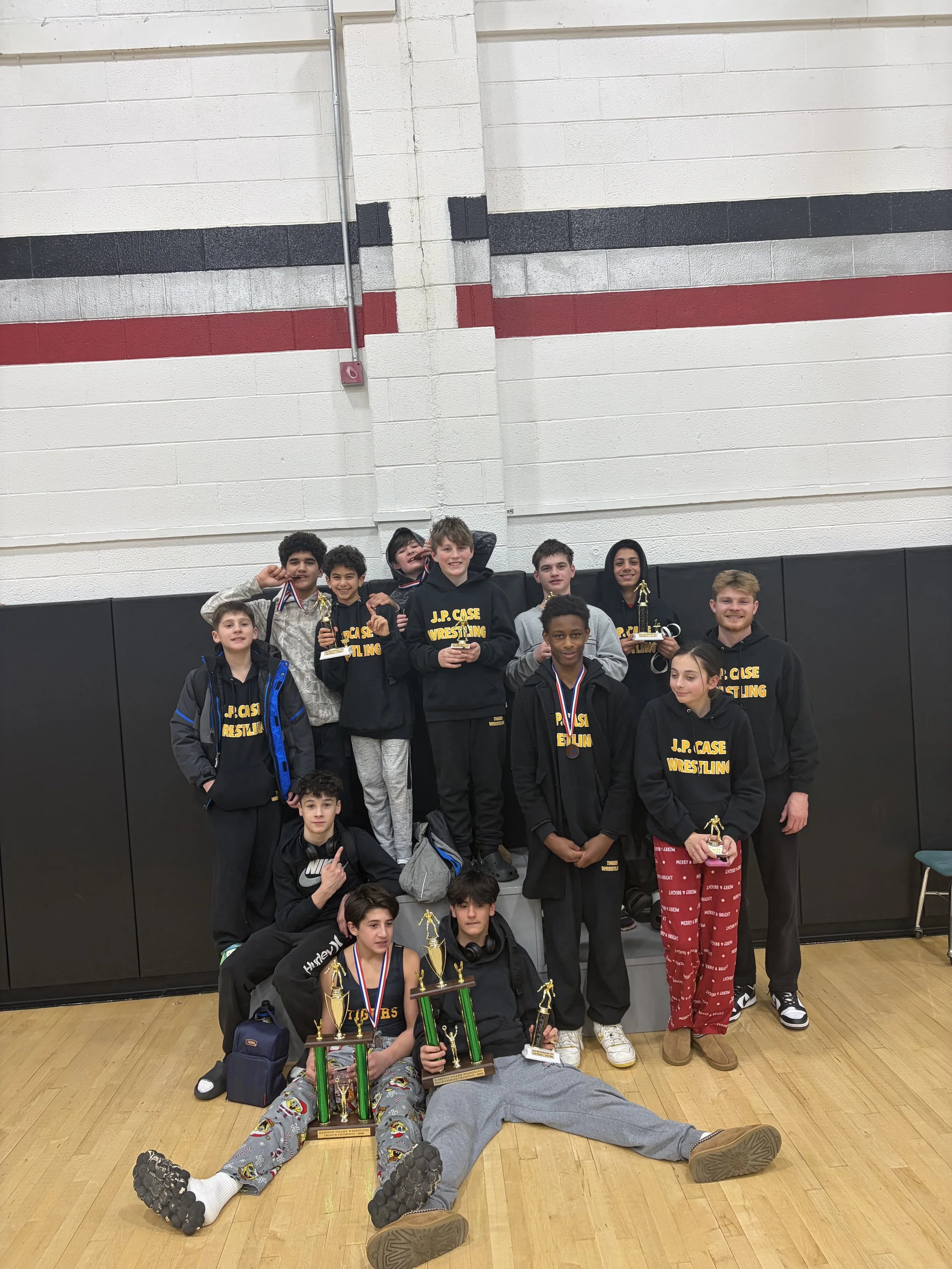 A group of young wrestlers, some holding trophies and medals, posing for a photo in a gymnasium with a white, black, and red wall in the background; some are standing, others are sitting or lying on the floor.