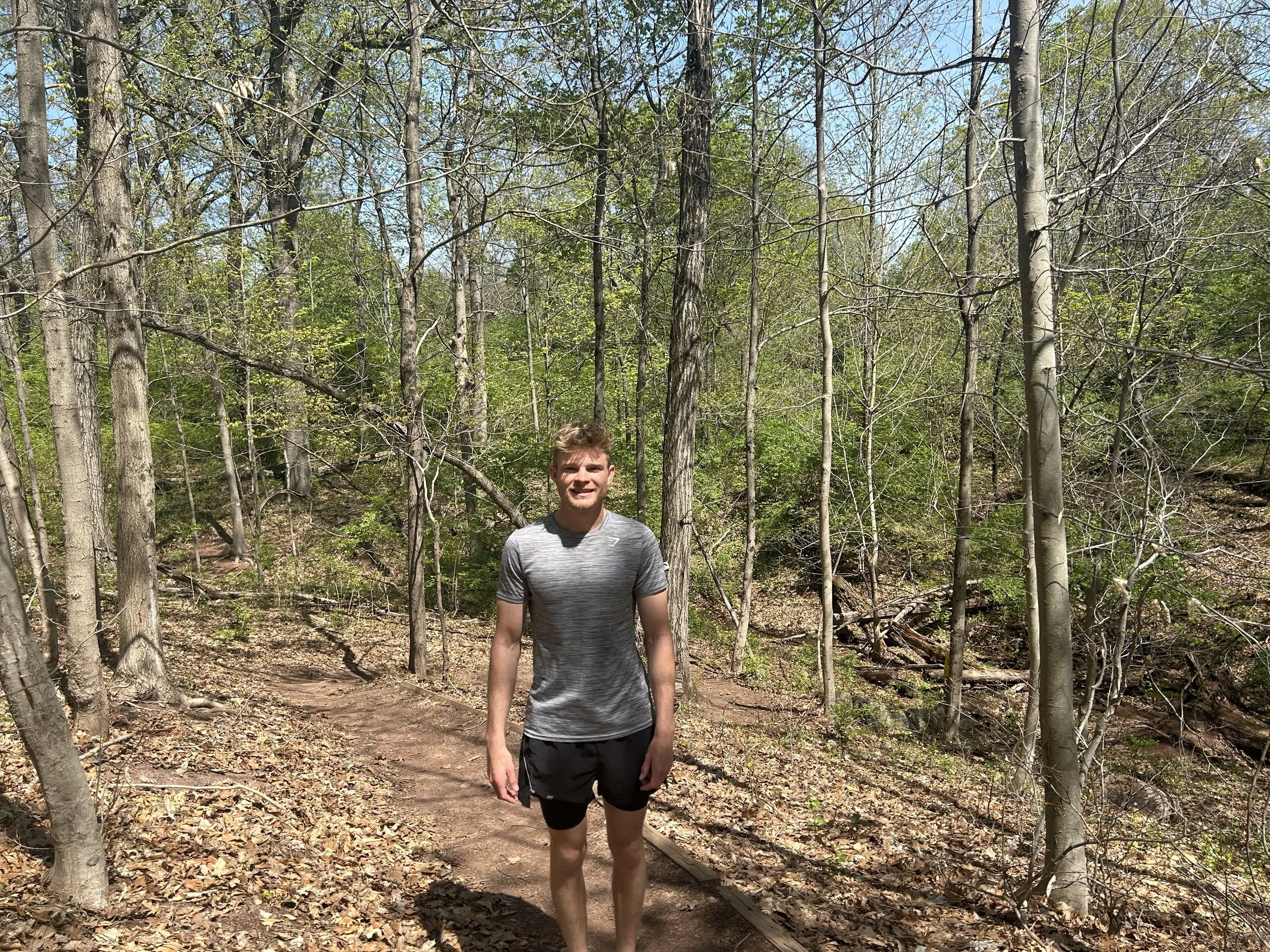 A young man with short curly hair, wearing a gray athletic shirt and black shorts, walking on a dirt trail in a wooded area with tall trees and green foliage.