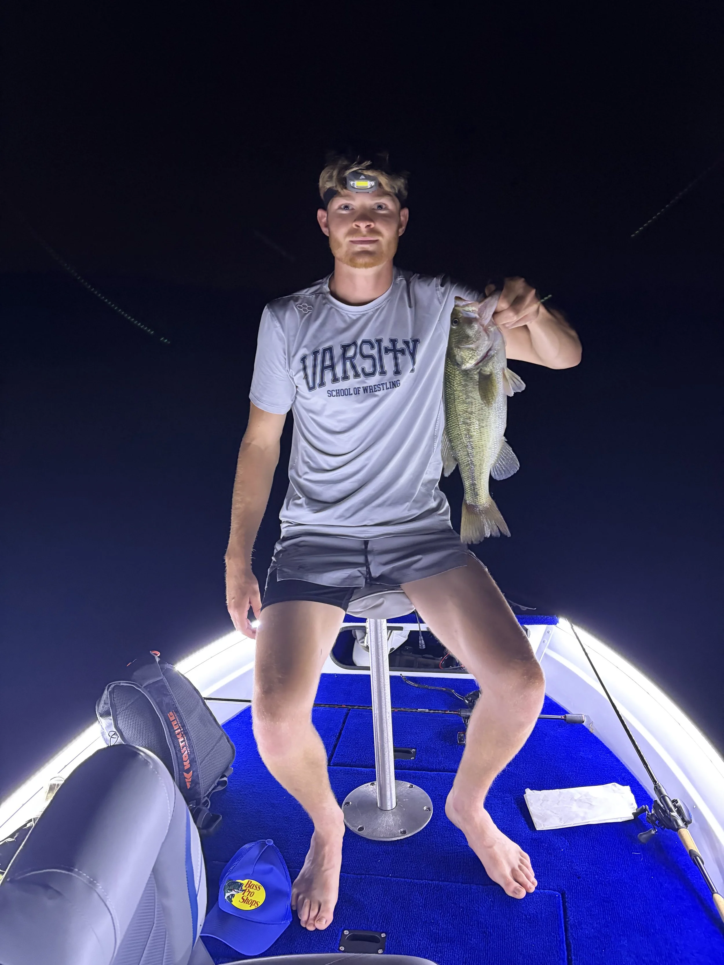 Young man with a headlamp holding a large fish on a boat at night, surrounded by fishing gear.