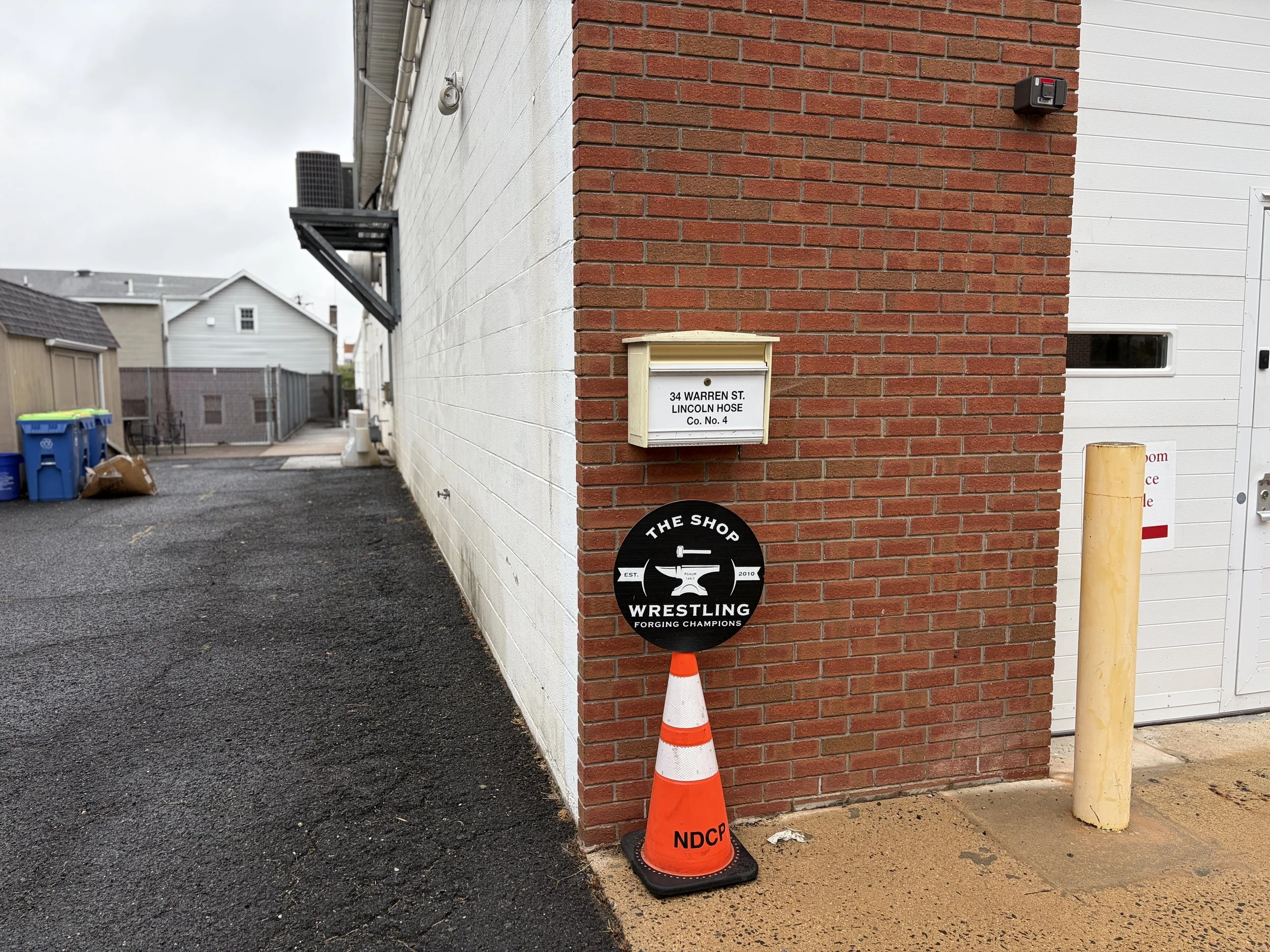 A corner of a building with a red brick wall and white siding, with a mailbox labeled "34 Warren St. Lincoln Hose Co. No. 4," a black wrestling sign, and orange traffic cones. There are trash bins and a fence visible in the background.