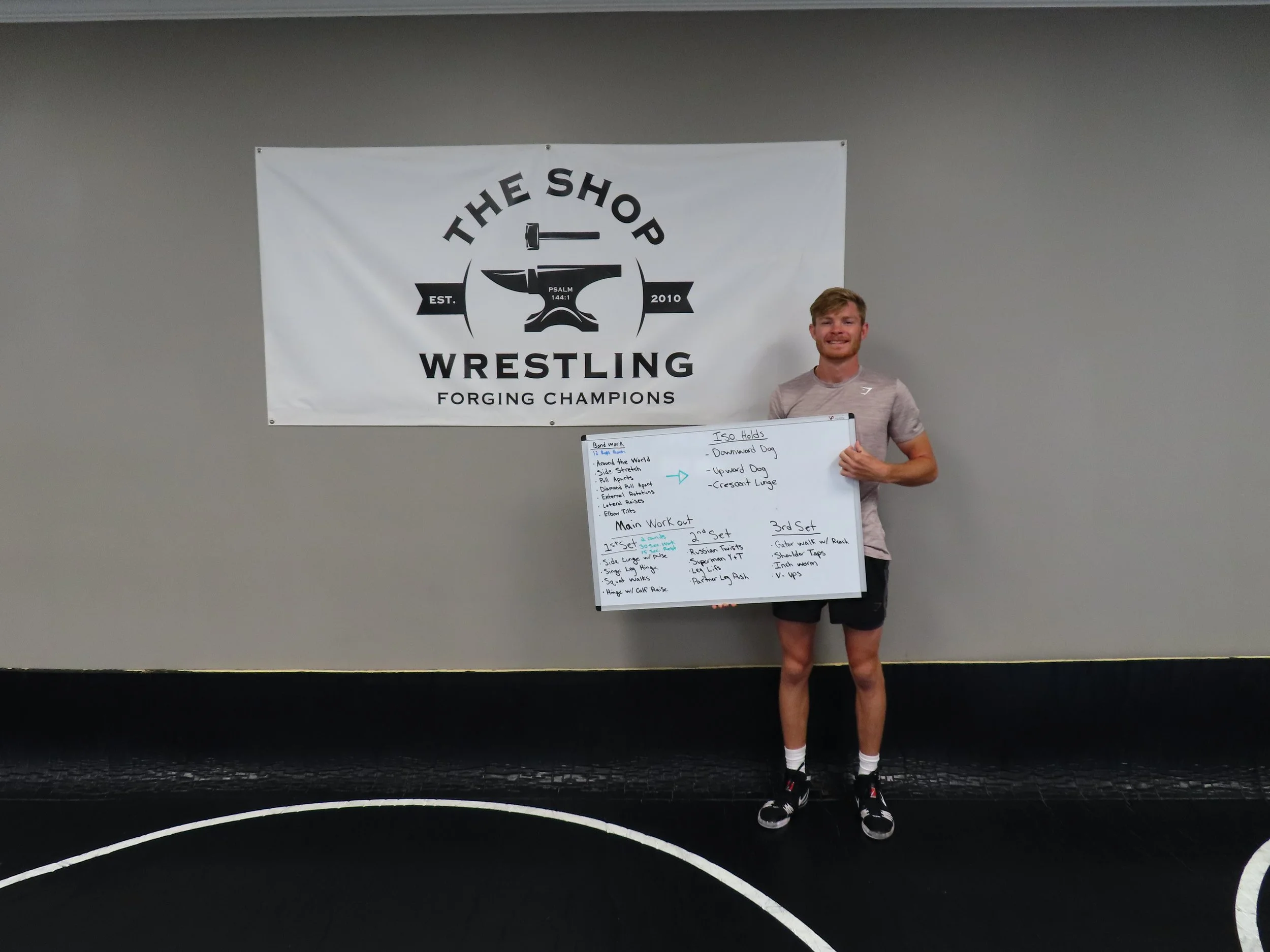 A young man standing in front of a whiteboard and a large banner that reads 'The Shop Wrestling Forging Champions' with a logo of a hammer, an anvil, and a banner with 'Est. 2010'. The young man is smiling, wearing athletic clothing, and holding a whiteboard with wedding planning notes.