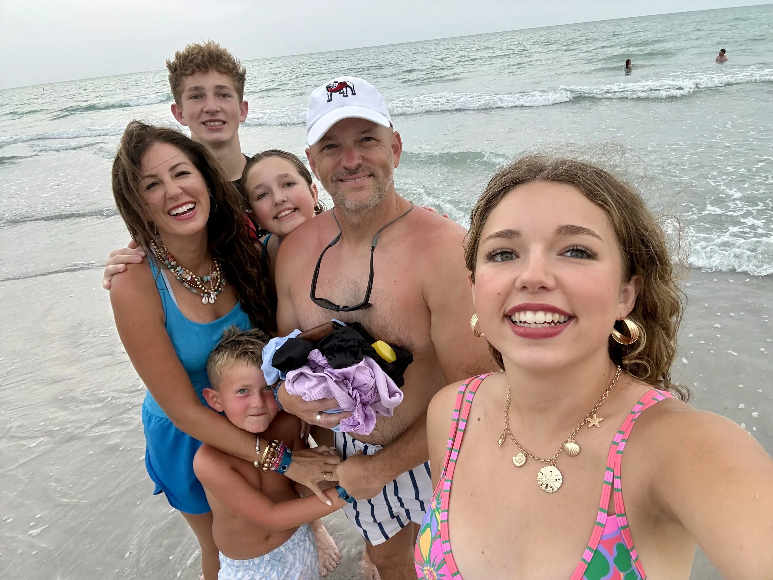 A family of six taking a selfie on the beach with the ocean in the background. The group includes two young girls, two boys, a woman, and a man, all smiling and enjoying their time.