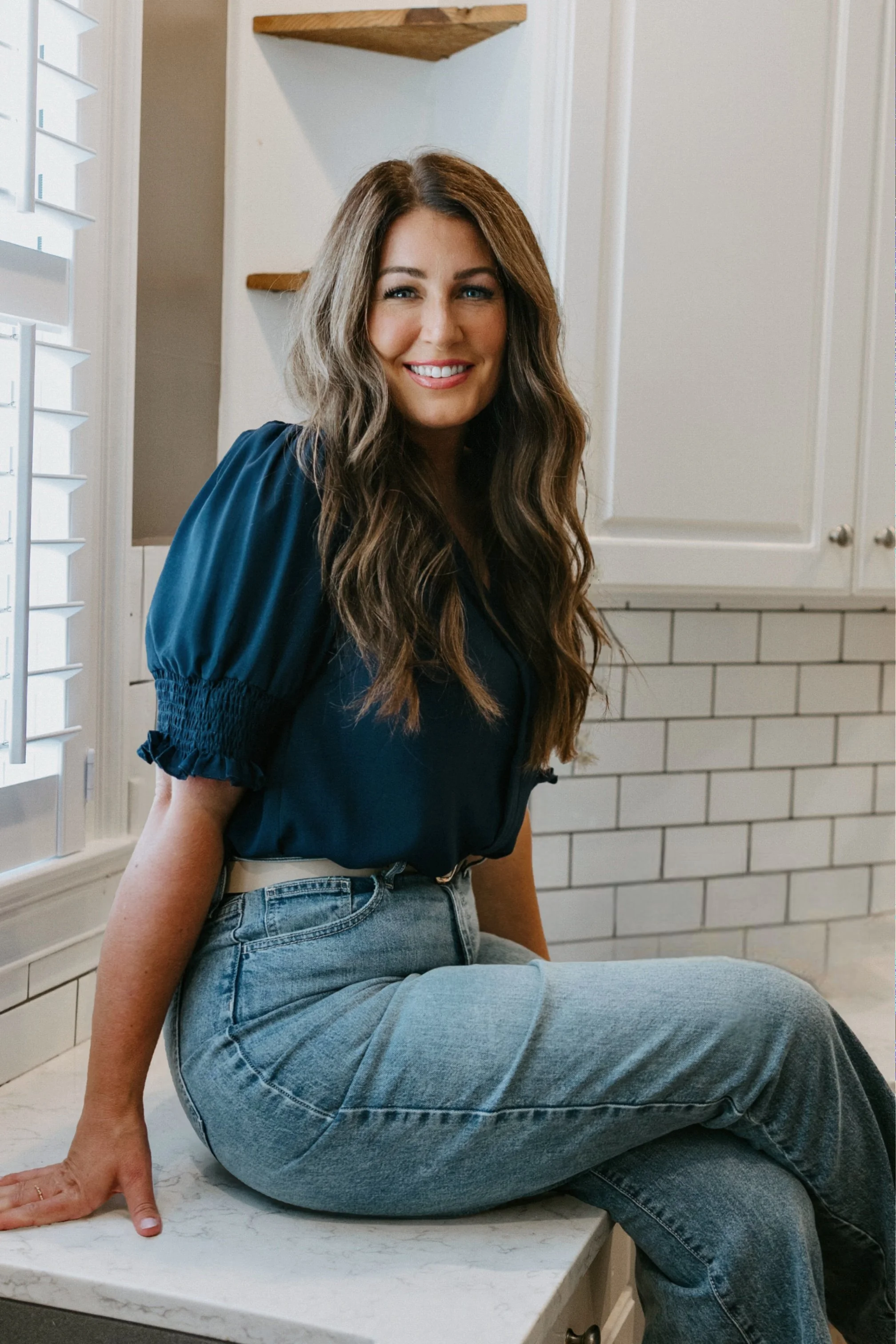 A woman with long wavy brown hair, wearing a dark blue blouse with puffed sleeves and light blue jeans, sits on a white kitchen countertop with white cabinets and a white tiled wall behind her. She has a bright smile and is looking at the camera.