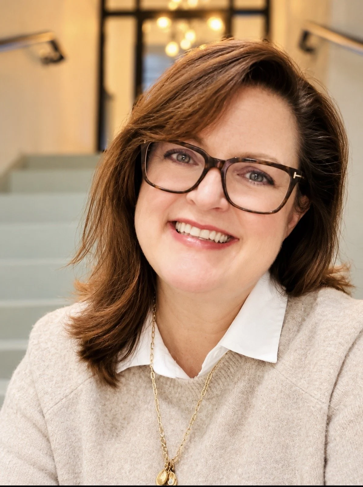 Smiling woman wearing glasses, a white collared shirt, and beige sweater in a home interior