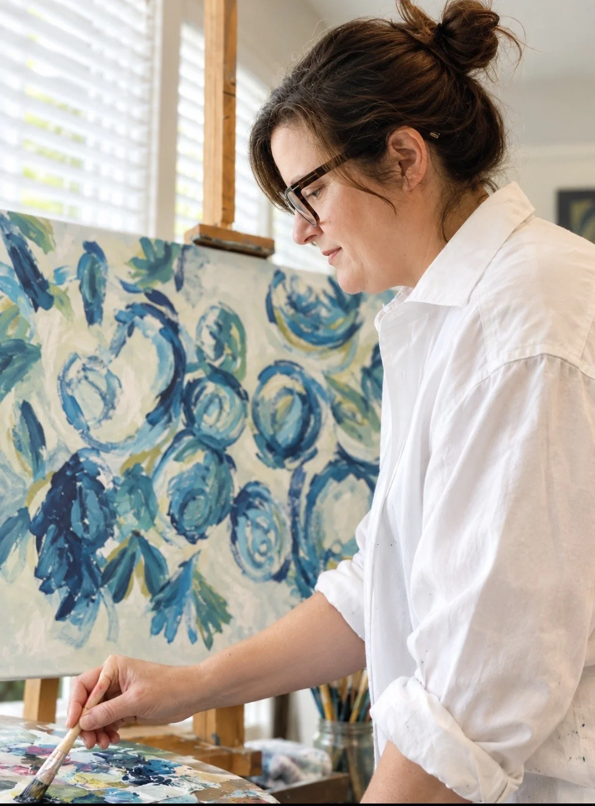 Woman wearing glasses and a white shirt painting a blue floral artwork on an easel
