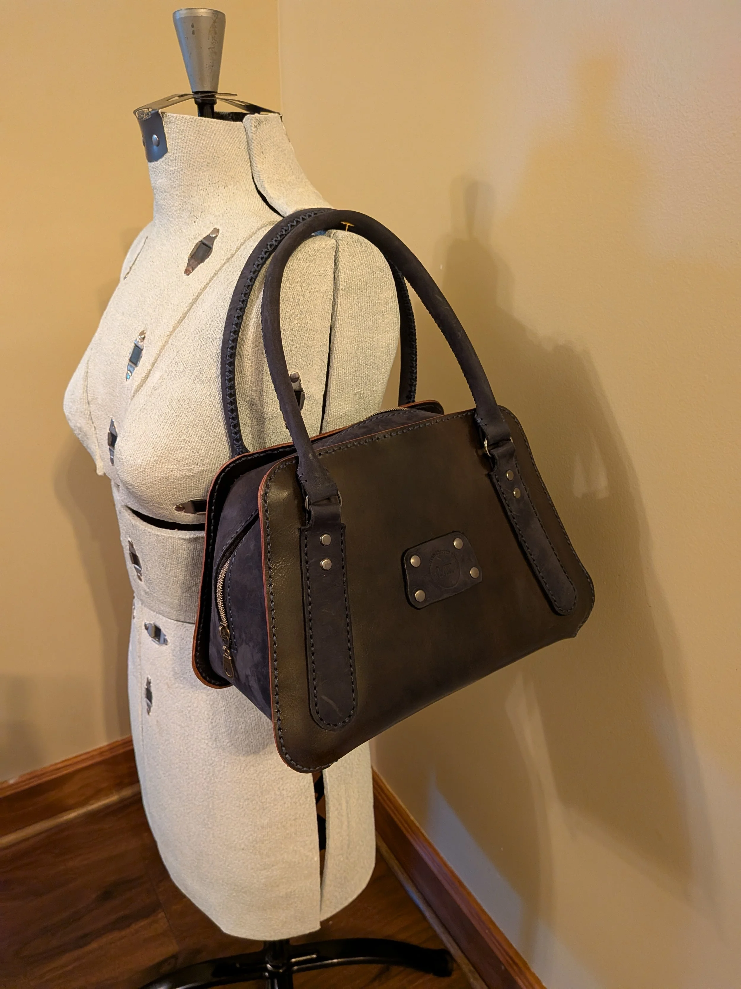 A dress form mannequin displaying a black leather handbag with brown accents, placed on a wooden floor next to a beige wall.