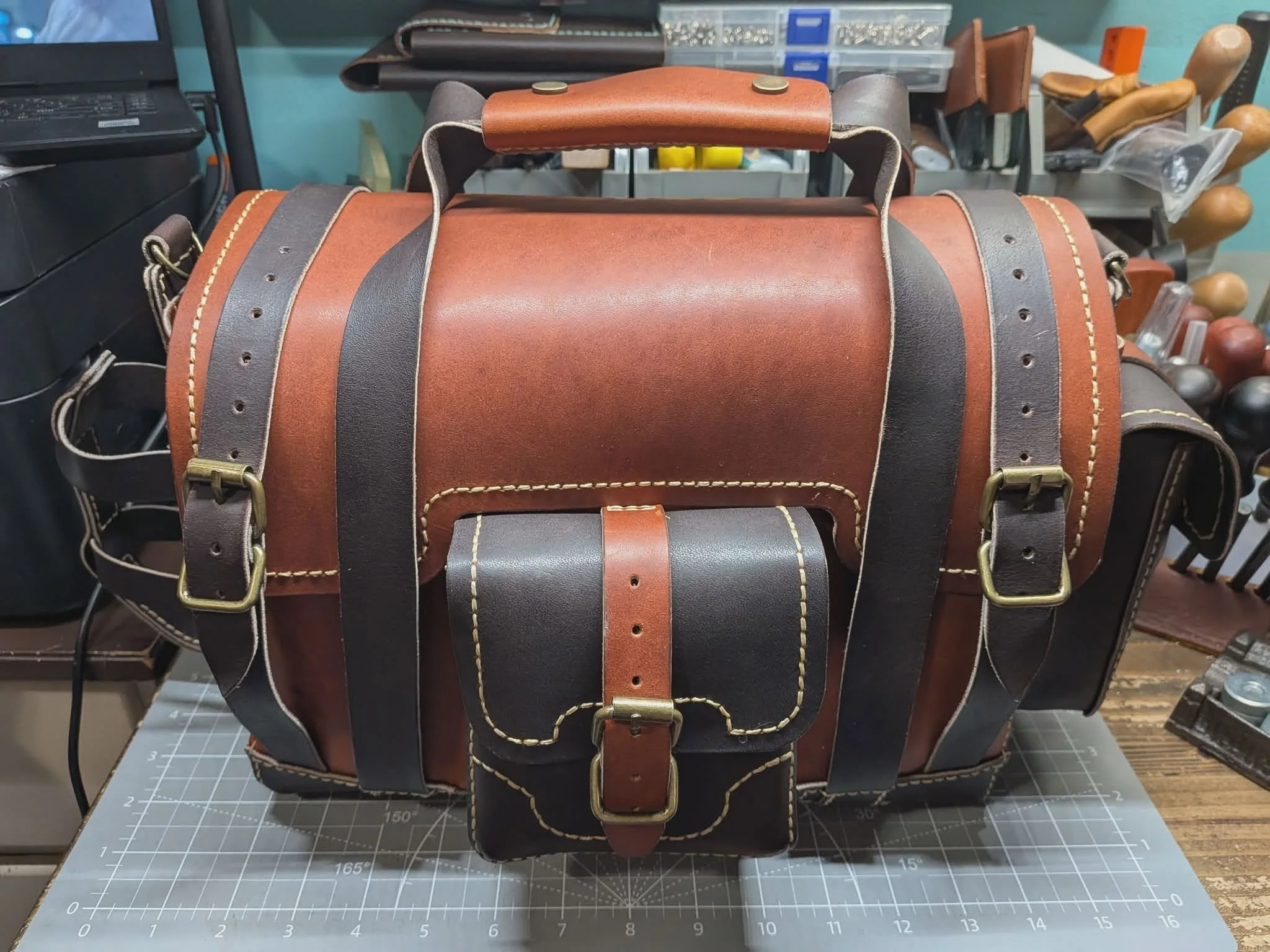 A leather tool bag with black and brown leather straps and brass buckles, placed on a workbench in a workshop with tools and supplies in the background.