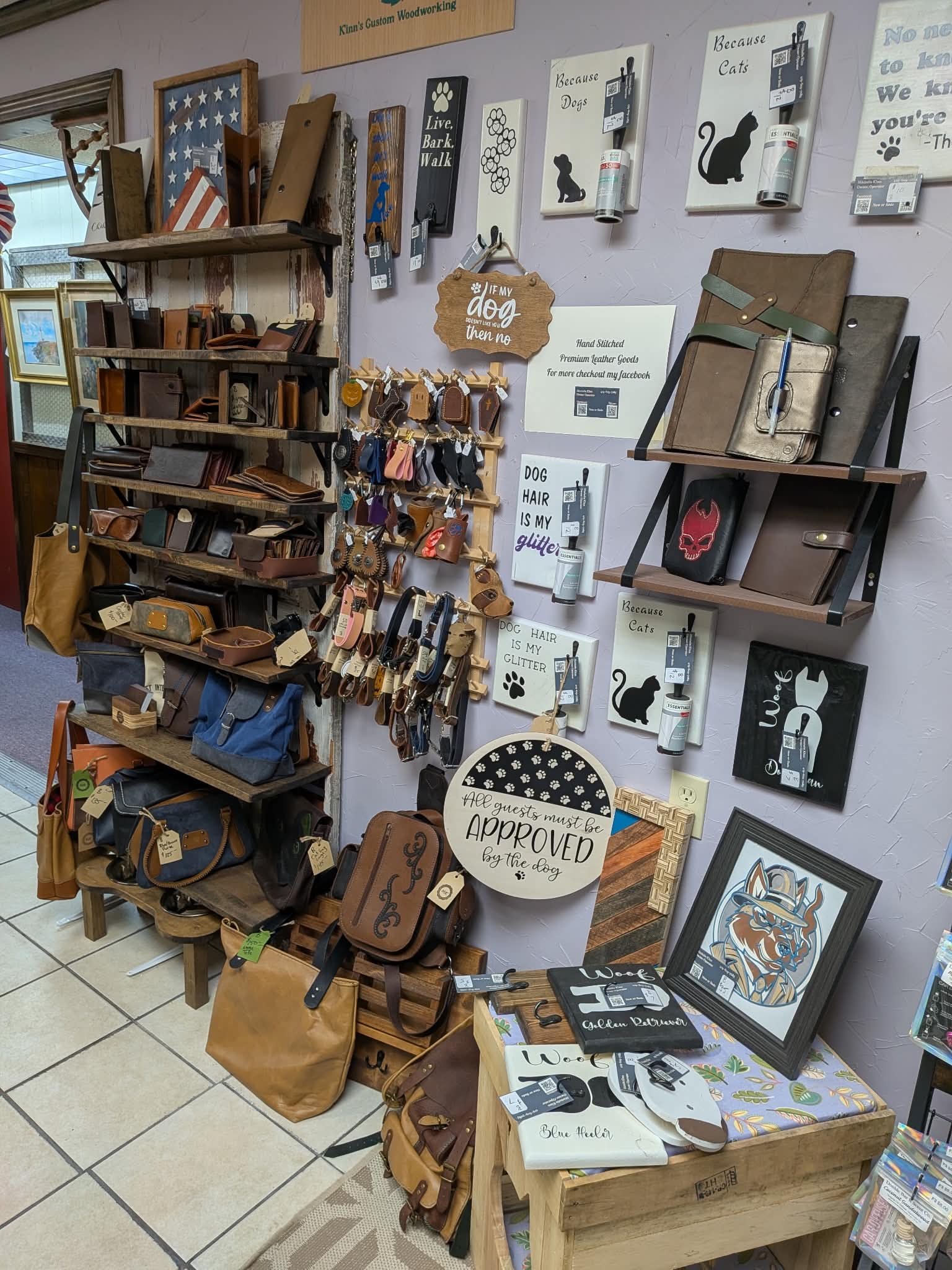 Display of leather goods including wallets, bags, and accessories on shelves and a table in a store. Wall decor includes signs with dog-themed phrases and artwork.