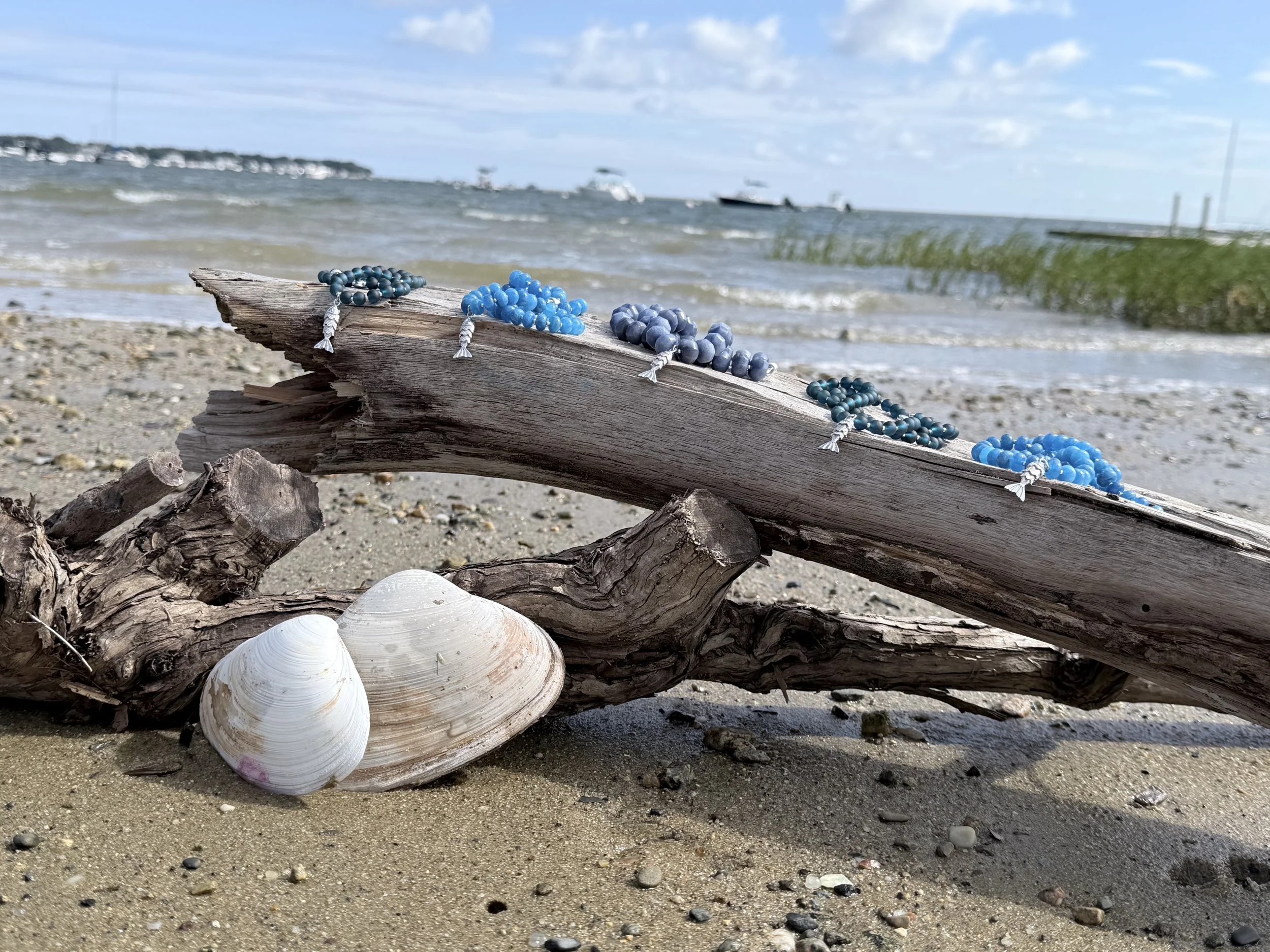 Colorful beaded necklaces and seashells arranged on driftwood on a sandy beach with water and boats in the background.