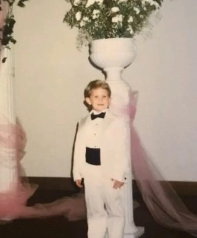 Young boy dressed in a white tuxedo with a black bow tie, standing next to a large white pedestal with a potted plant on top, at an indoor event.