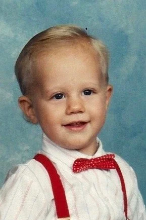 A young boy with blond hair, wearing a white shirt with red suspenders and a red polka dot bow tie, smiling against a blue background.