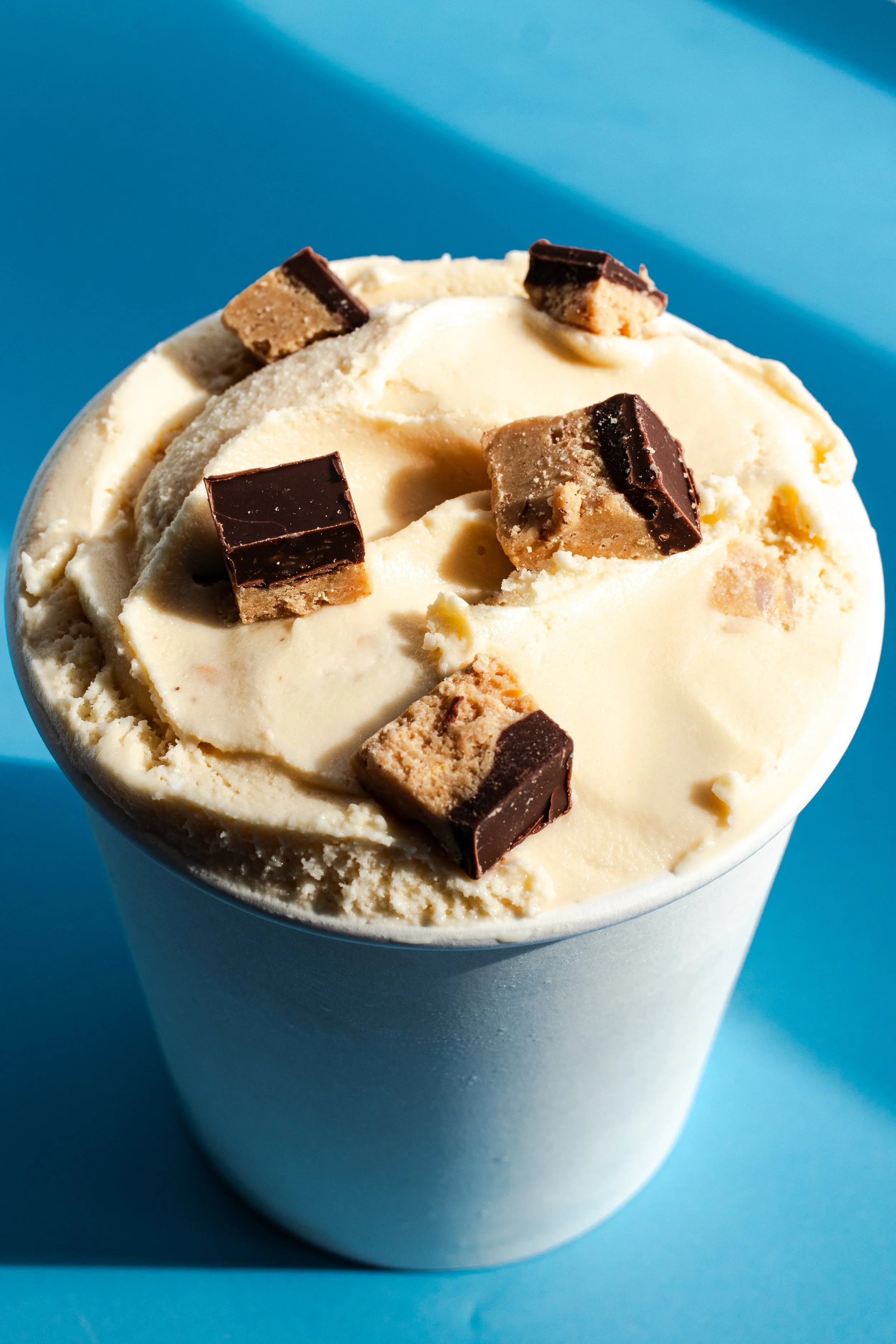 A cup of vanilla ice cream topped with chocolate and peanut butter chunk pieces on a blue background.