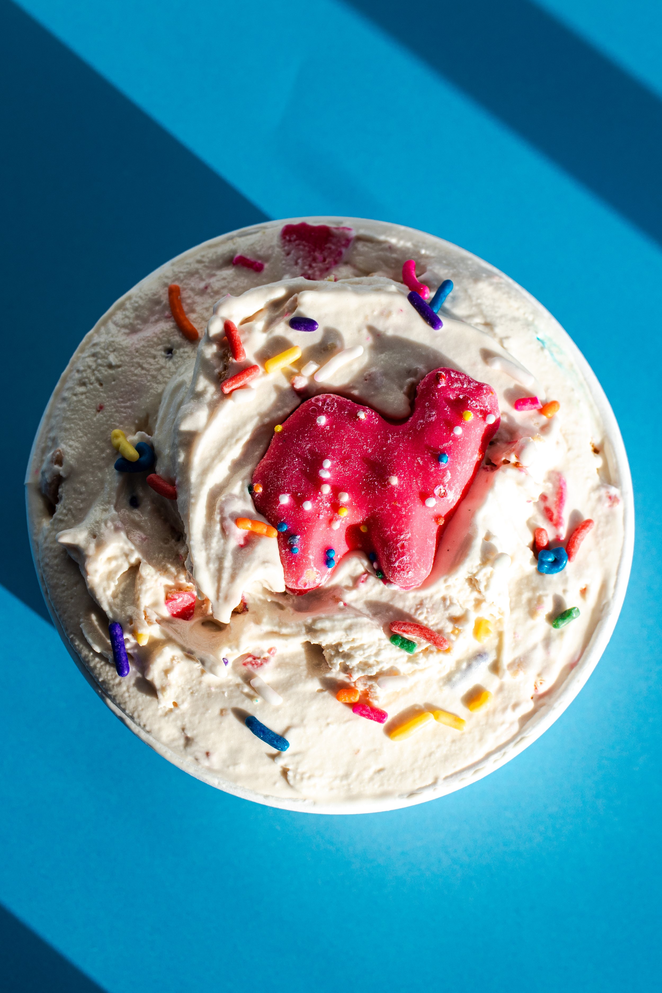 A close-up of a cup of vanilla ice cream topped with rainbow sprinkles, a red gummy candy, and pink syrup, set against a blue background.