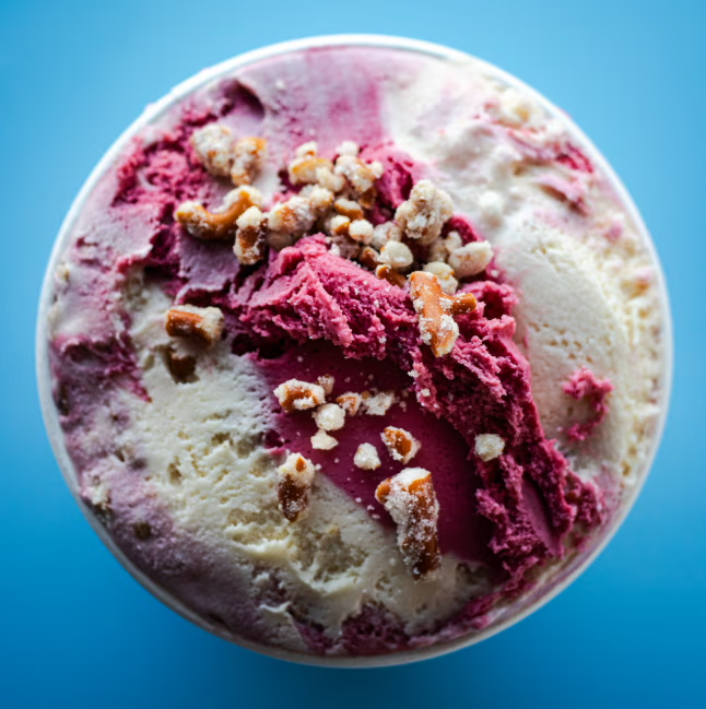 A close-up of a bowl of ice cream with pink and white swirls, topped with caramelized nuts, against a blue background.