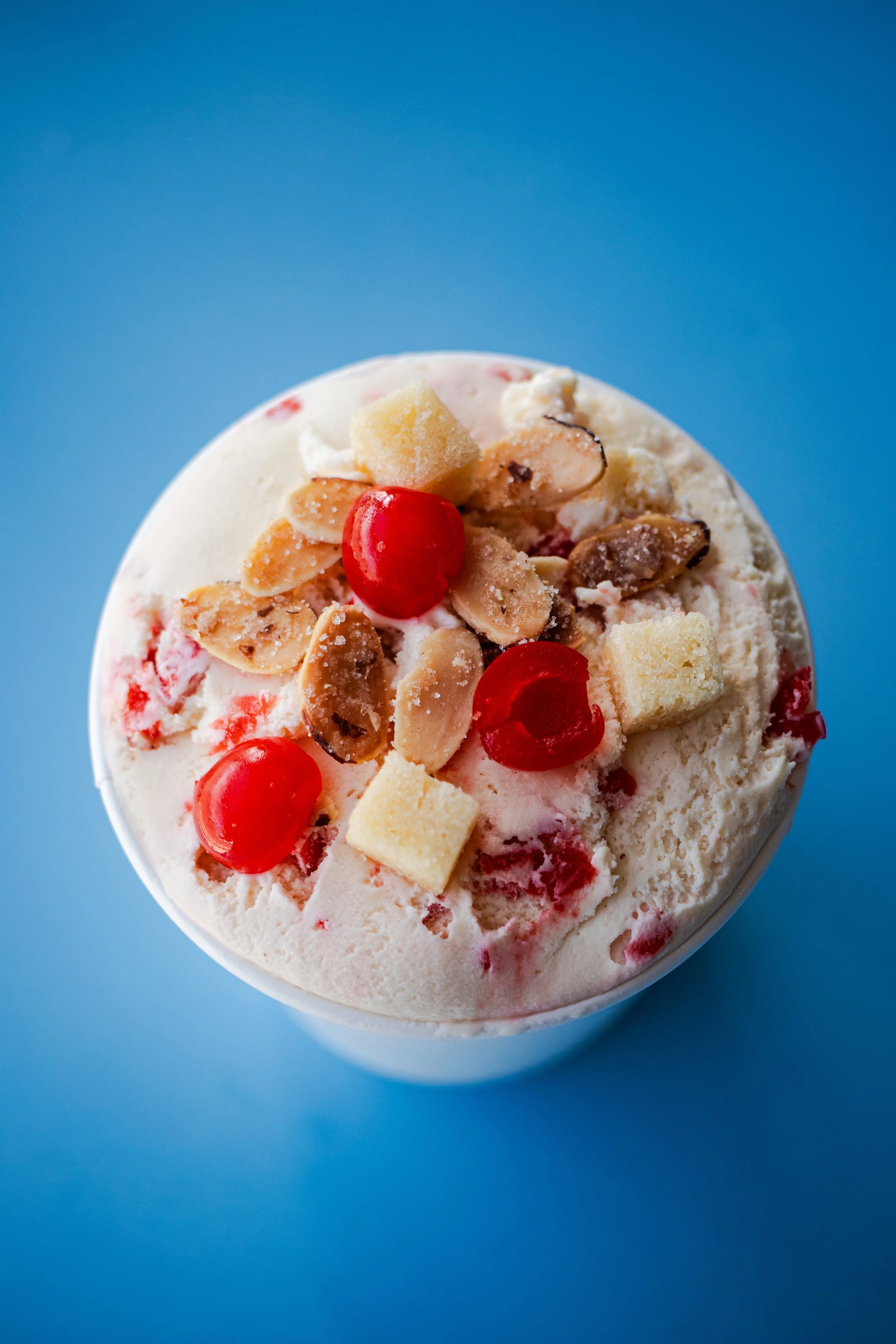 Close-up of a scoop of ice cream with red cherries, chopped nuts, and pieces of cake on top, set against a blue background.