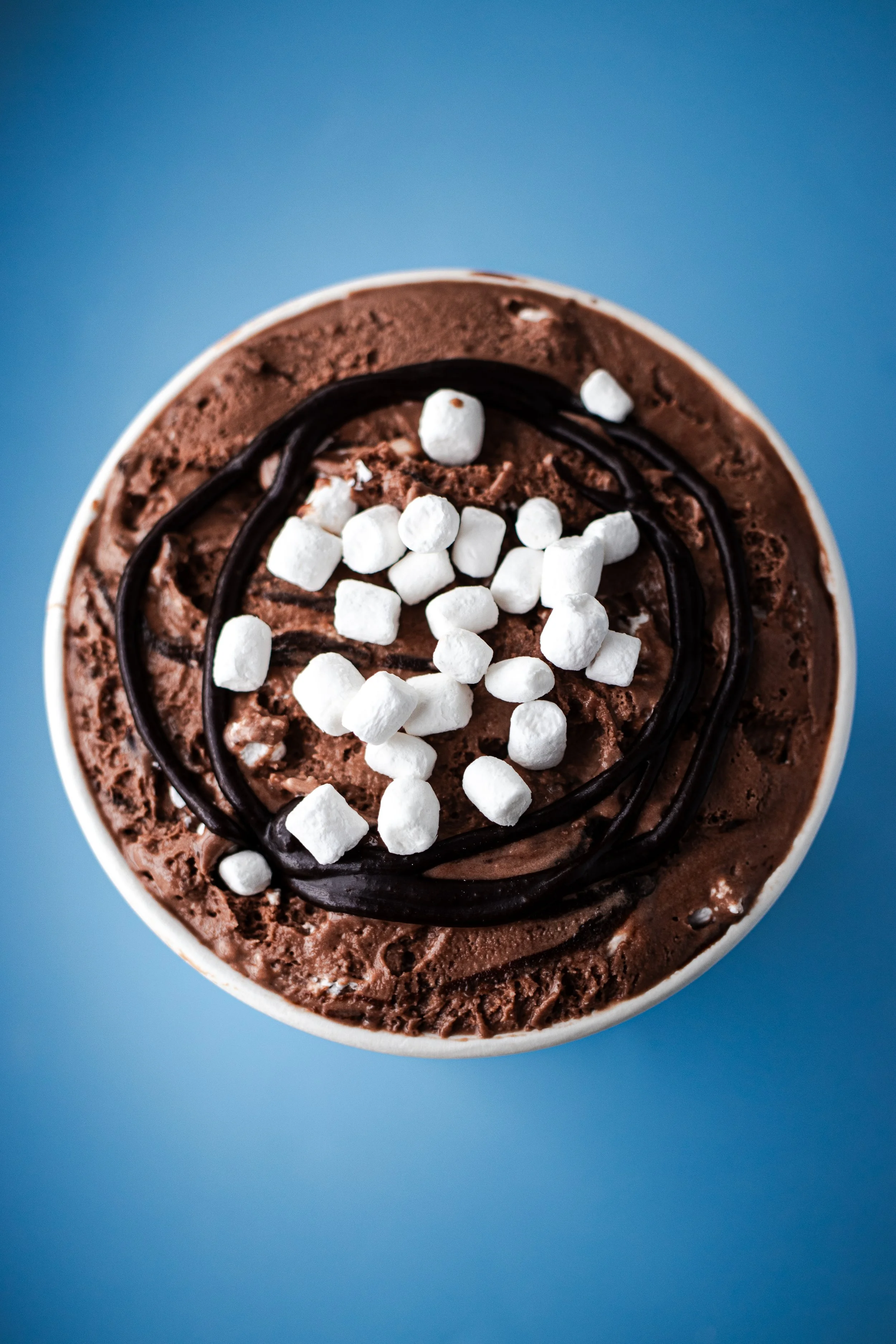 Chocolate dessert topped with mini marshmallows and a drizzle of chocolate sauce, against a blue background.