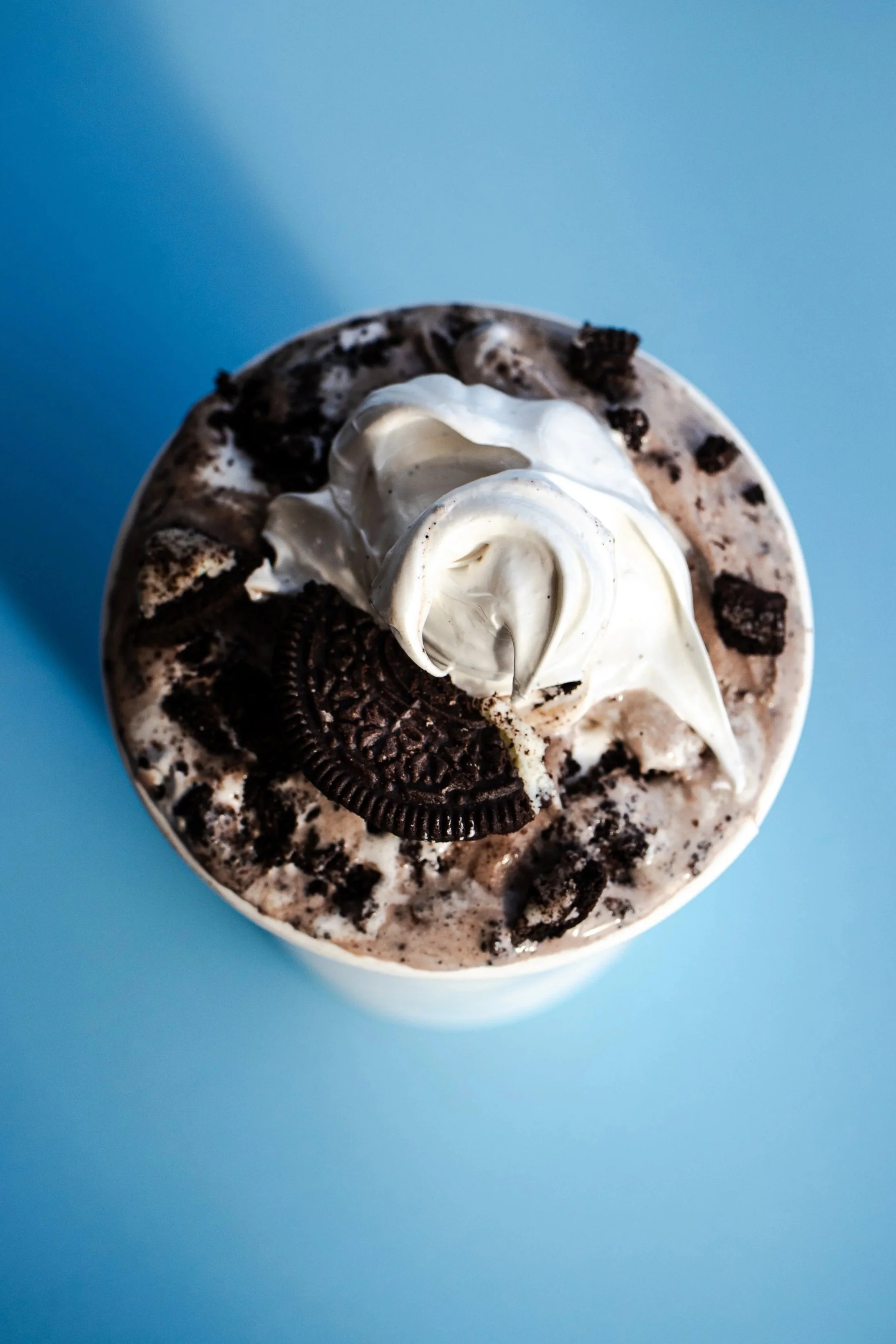 Close-up of a cookies and cream flavored ice cream topped with whipped cream, crushed cookies, and whole Oreo cookies, against a blue background.