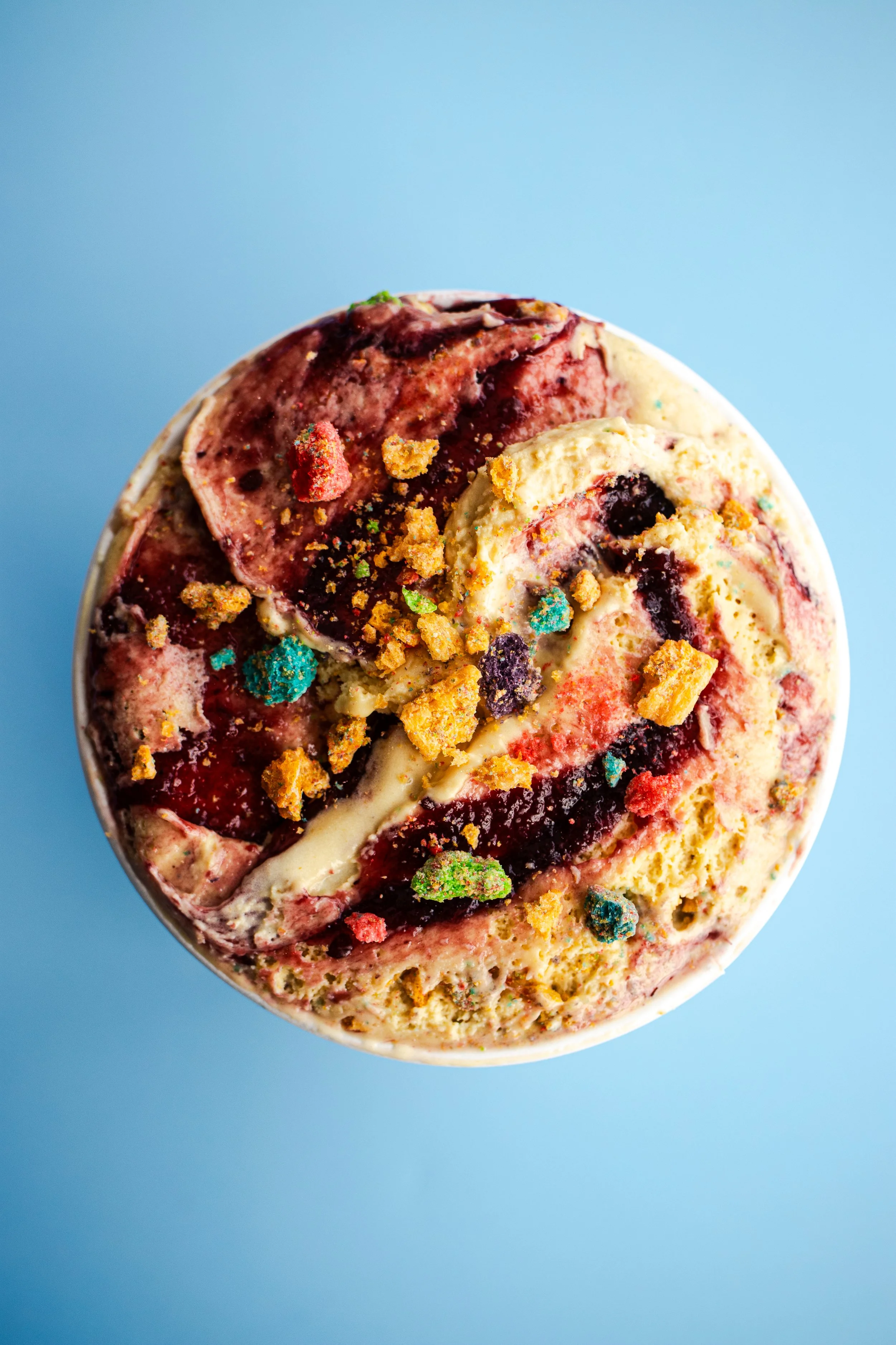 Close-up of a colorful ice cream swirl topped with colorful sprinkles and crushed cookies, set against a blue background.