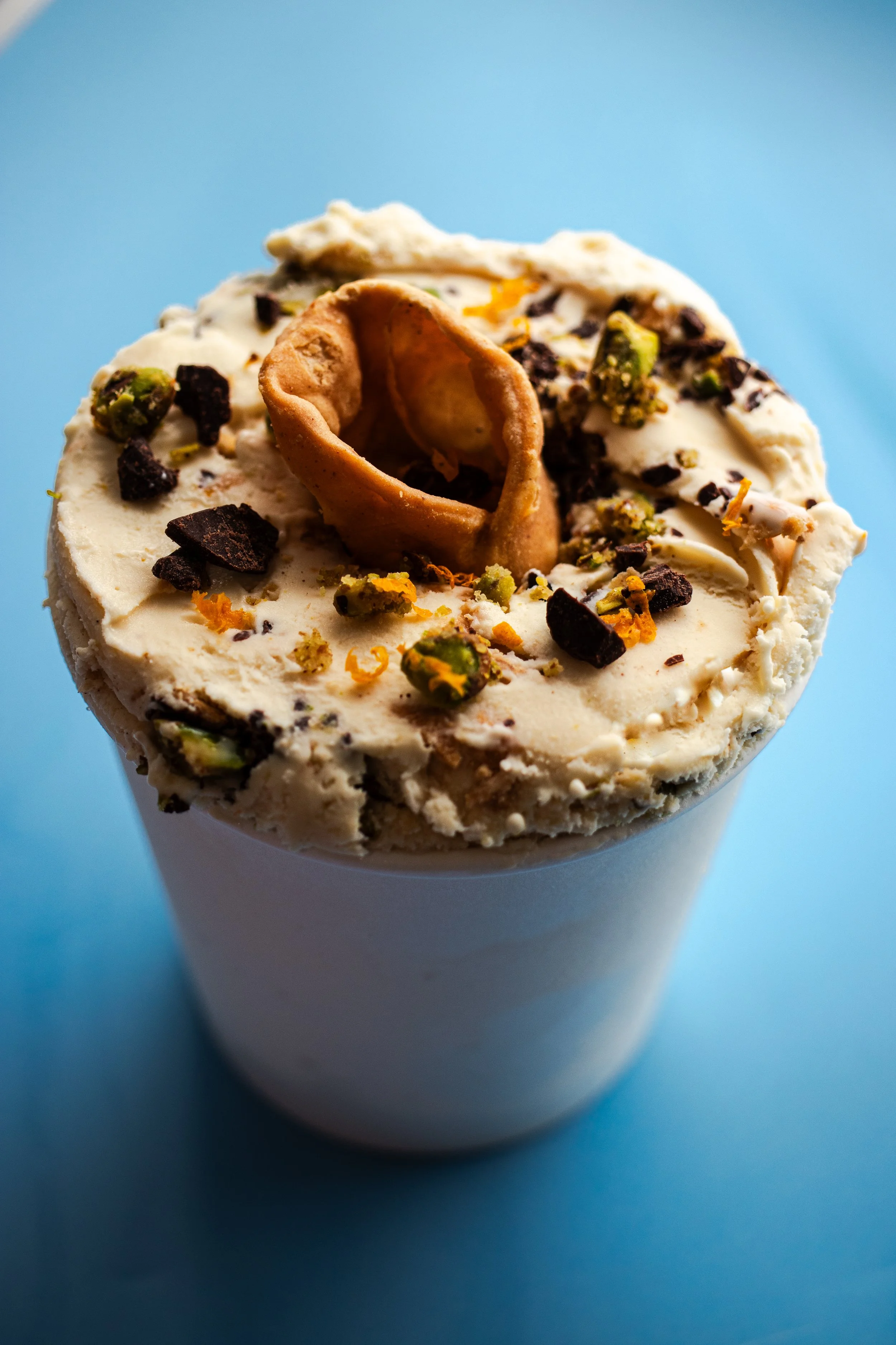 Close-up of a vanilla ice cream cup topped with chocolate shavings, chopped pistachios, and a rolled cookie on top, against a blue background.
