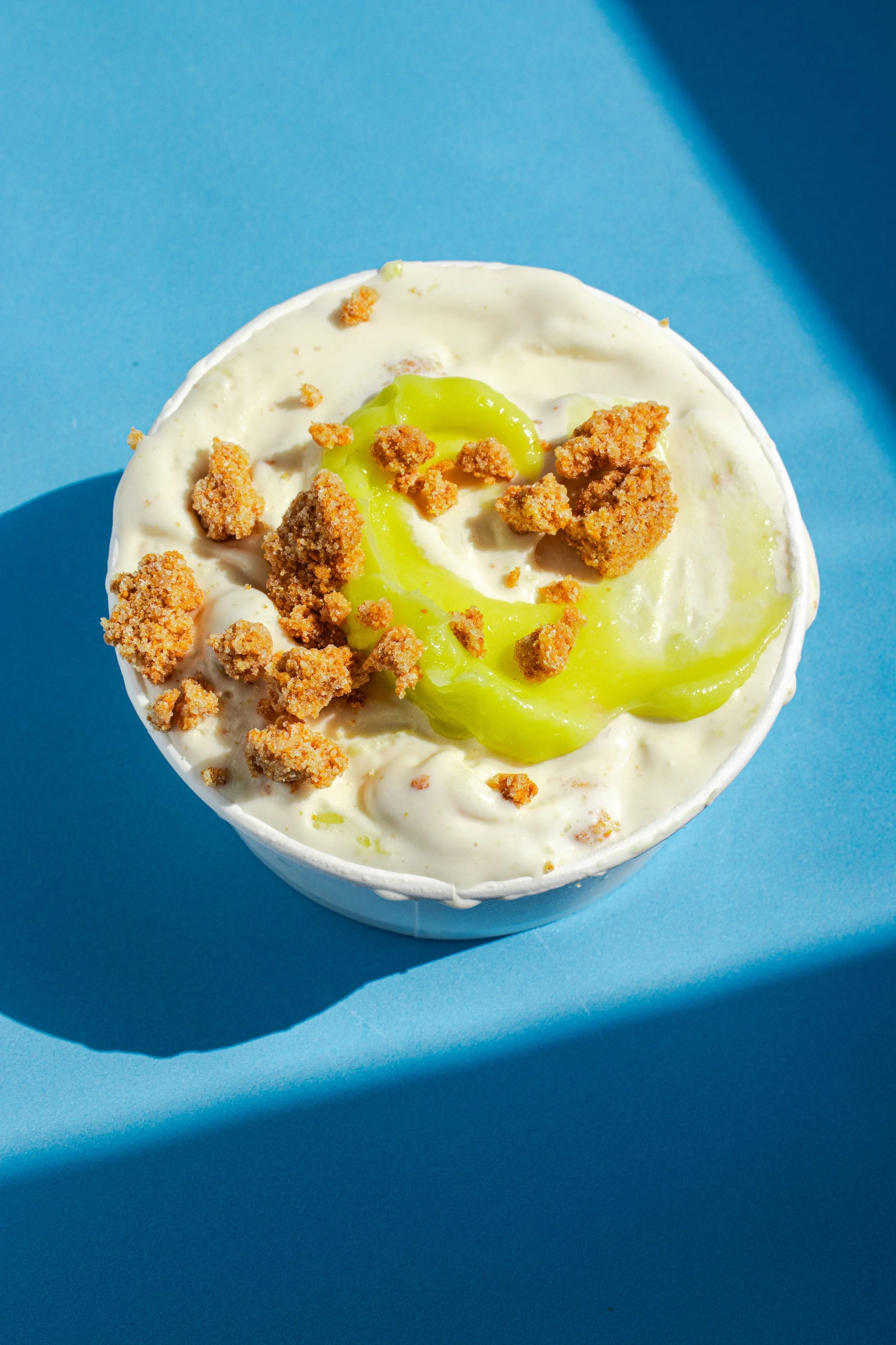 Close-up of a cup of ice cream with green syrup and crumbled cookies on top, on a blue background.