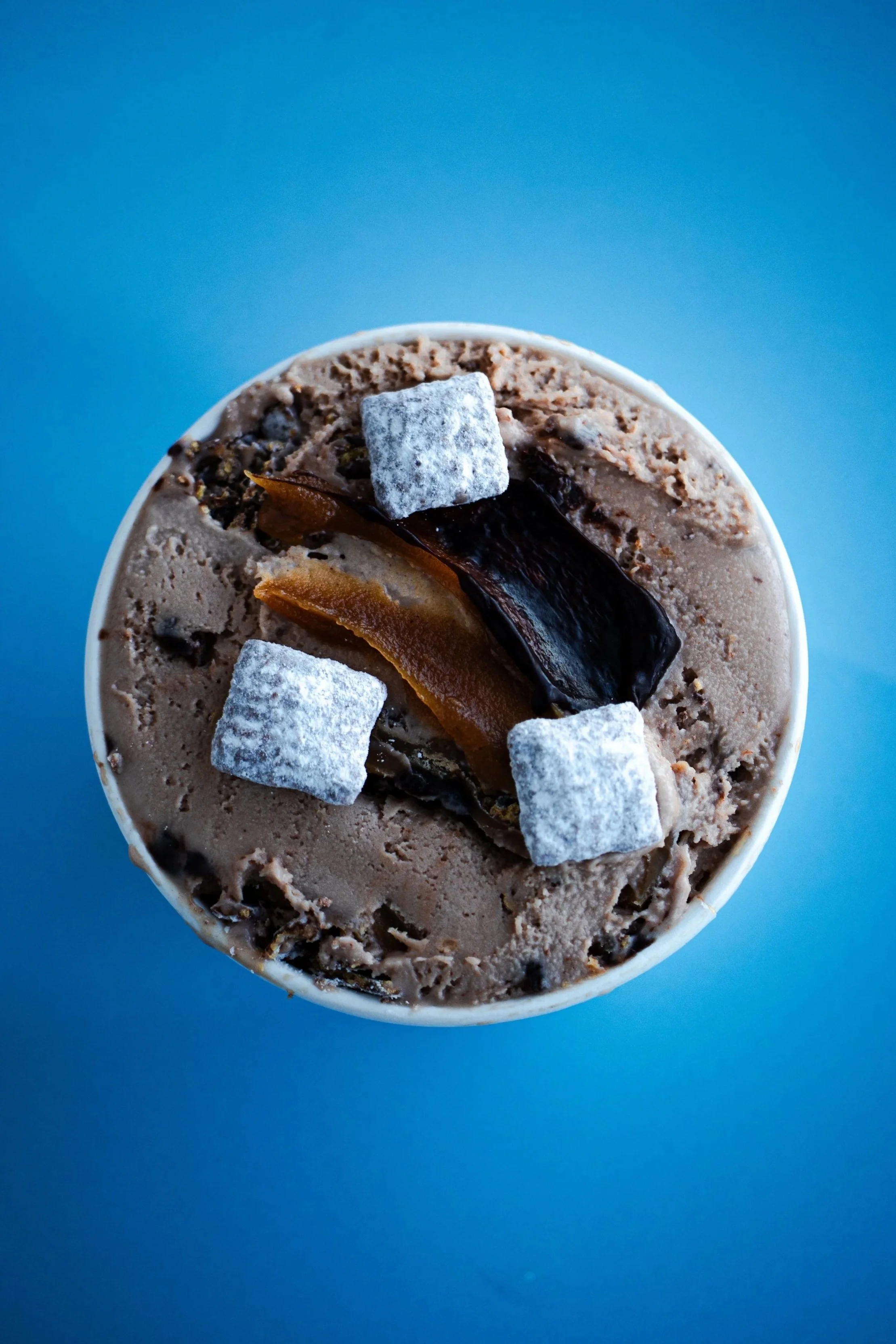 Close-up of a pint of ice cream cup topped with muddy buddies and chocolate against a bright blue background.