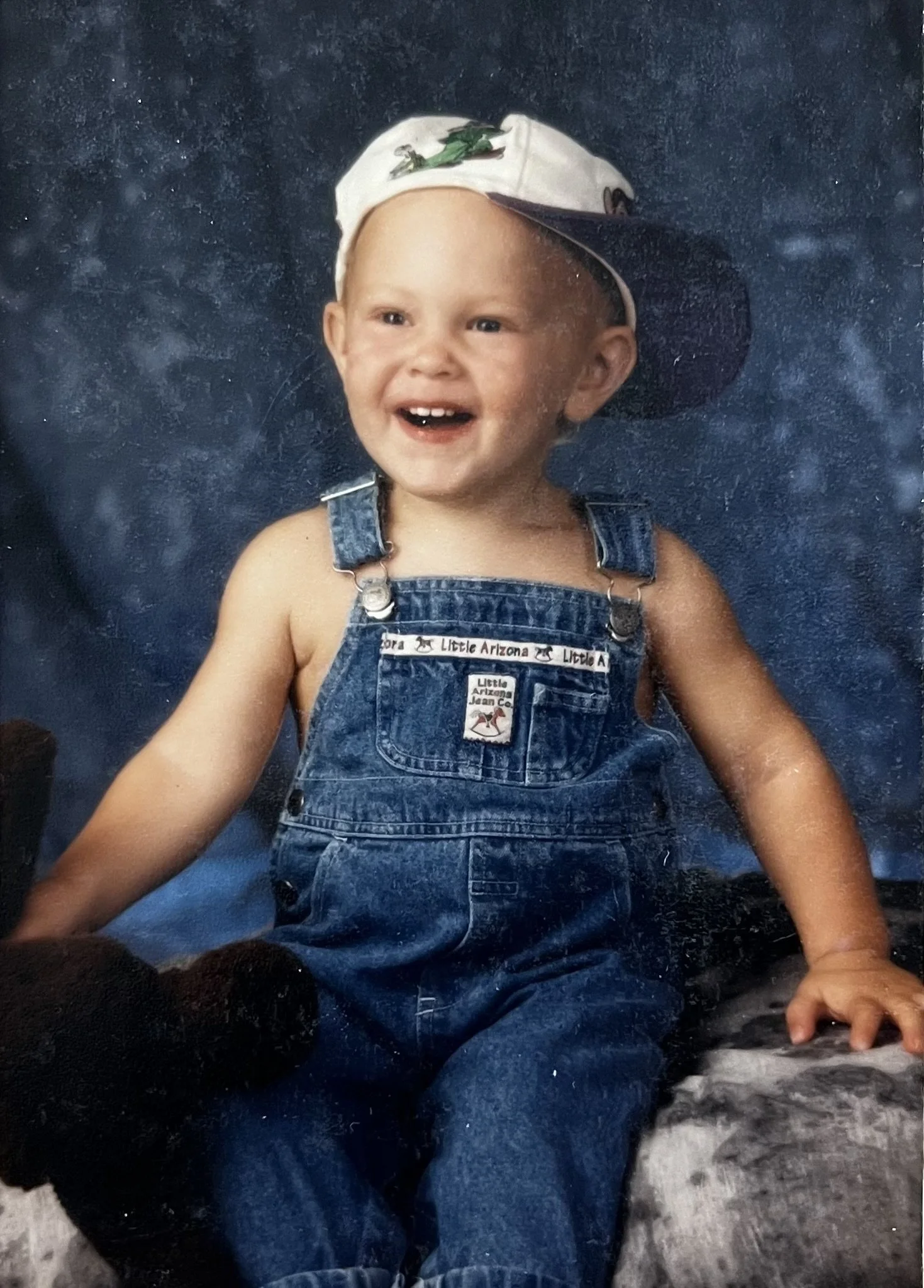 A young child wearing a white baseball cap with a lizard graphic and denim overalls is sitting on a large rock next to a flowing stream, smiling happily.