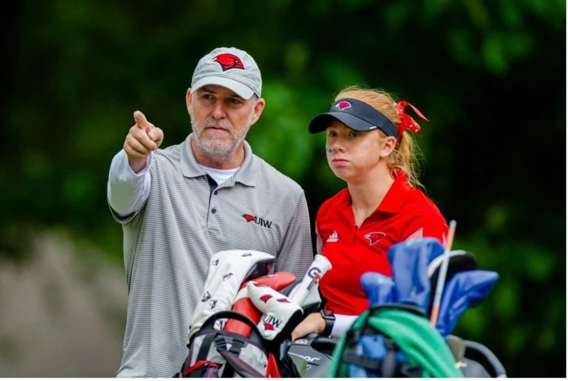 A man and a young woman, both wearing golf attire, are on a golf course surrounded by greenery. The man is pointing, and the woman looks attentive, holding a golf club with golf bags nearby.