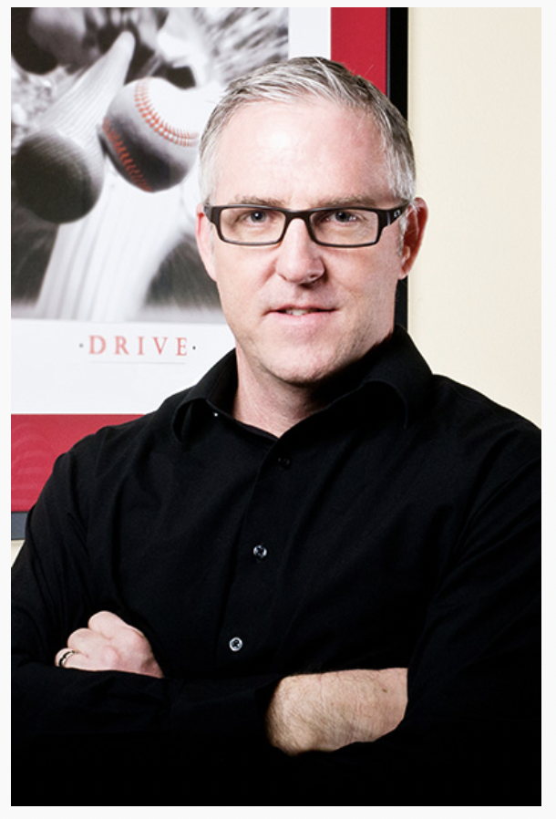A man with short gray hair and glasses, wearing a black shirt, standing with arms crossed in front of a baseball-themed poster.