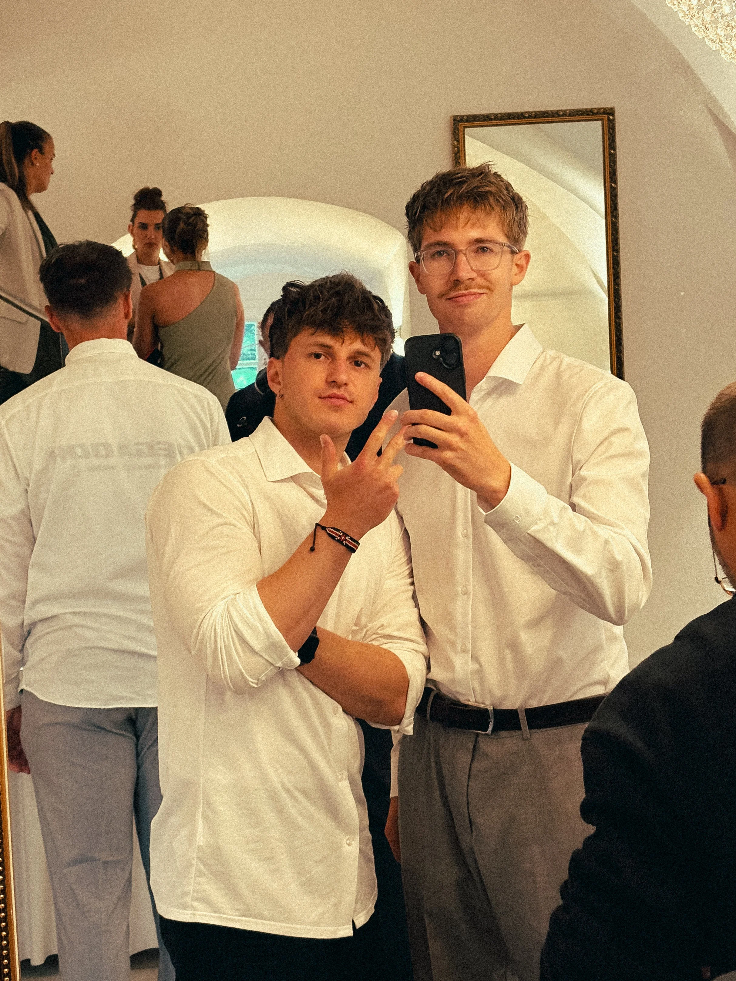 Zwei junge Männer nehmen im Spiegelselfie auf, umgeben von mehreren Personen in einem Raum, vermutlich bei einer Veranstaltung.