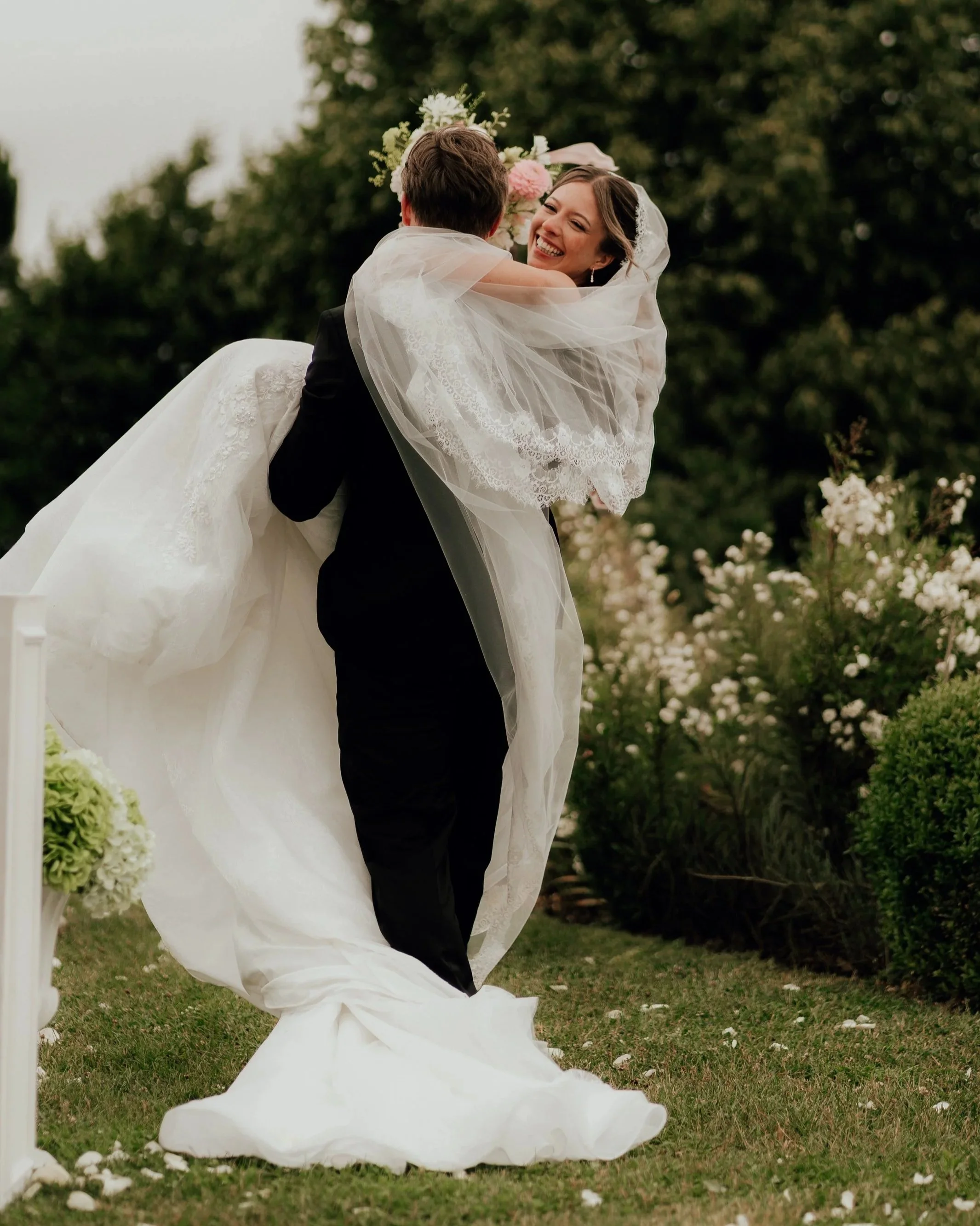 Ein Brautpaar liebt sich bei einer Hochzeit im Freien, die Frau trägt ein weißes Brautkleid und eine Schleier, der Mann trägt einen schwarzen Anzug. Sie lachen sich an und an einem Baum sind Blumenblumen zu sehen.