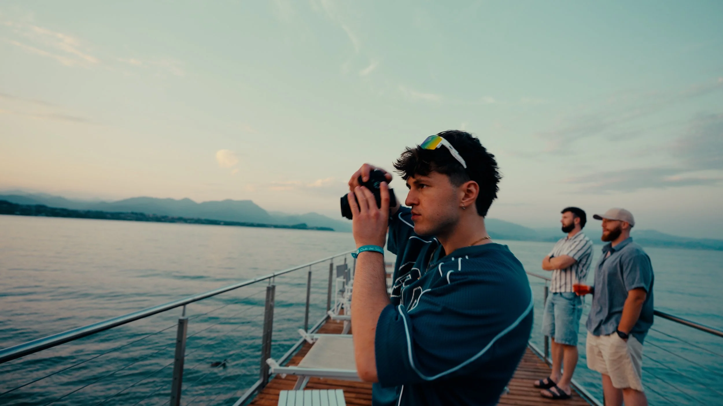 Junge Person mit dunklen Haaren und Brille auf dem Kopf, die eine Kamera auf einem Pier am Wasser benutzt, mit zwei Männern im Hintergrund, die am Wasser stehen, bei Sonnenuntergang.