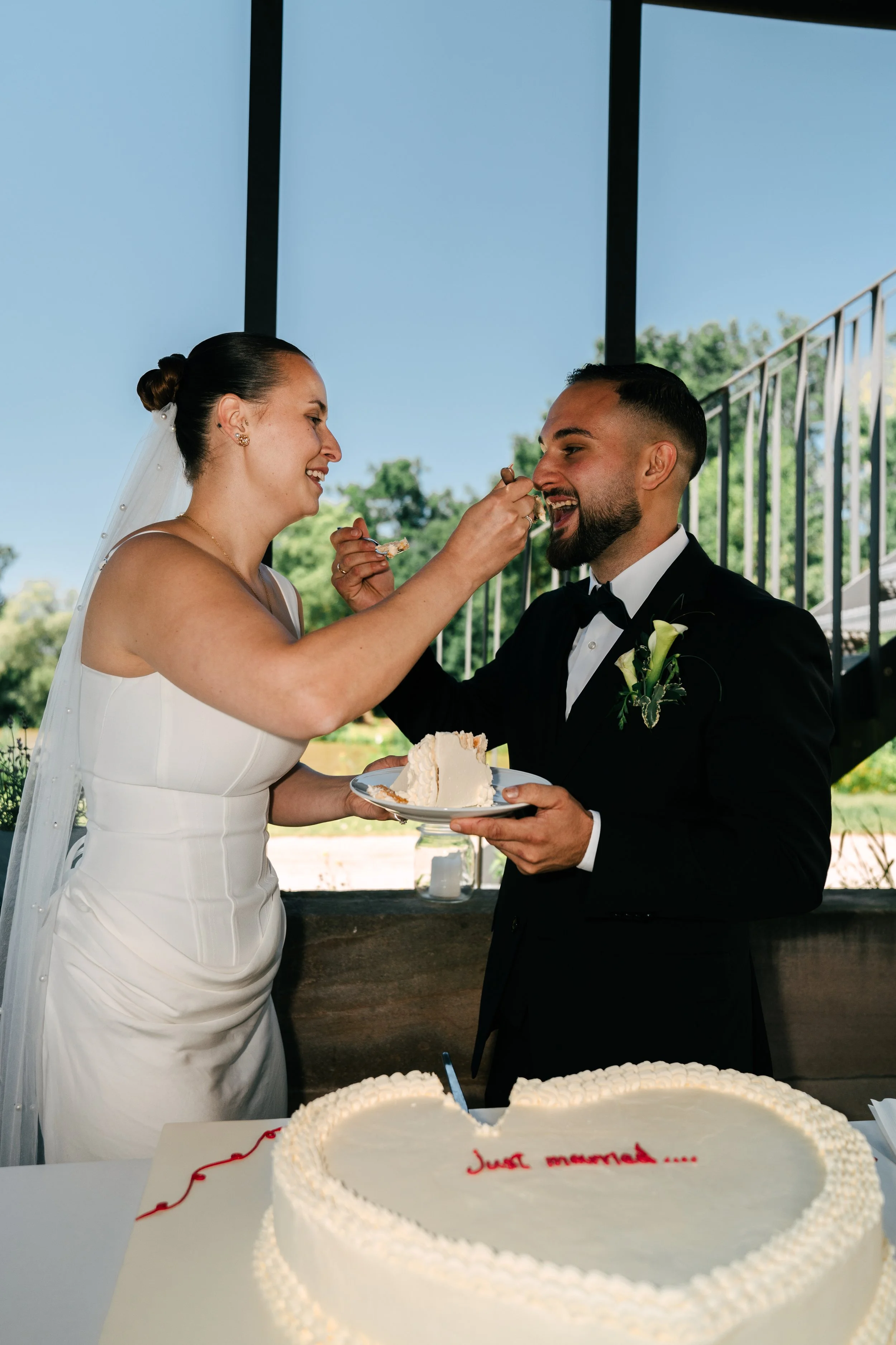 Ein Brautpaar beim Anschneiden der Hochzeitstorte, die Braut trägt ein weißes Kleid und eine Schleier, der Bräutigam ein schwarzes Anzug mit Fliege, im Hintergrund sind Bäume und eine Treppe zu sehen, im Vordergrund ein Herz-förmiger Kuchen mit der Aufschrift "Just married".