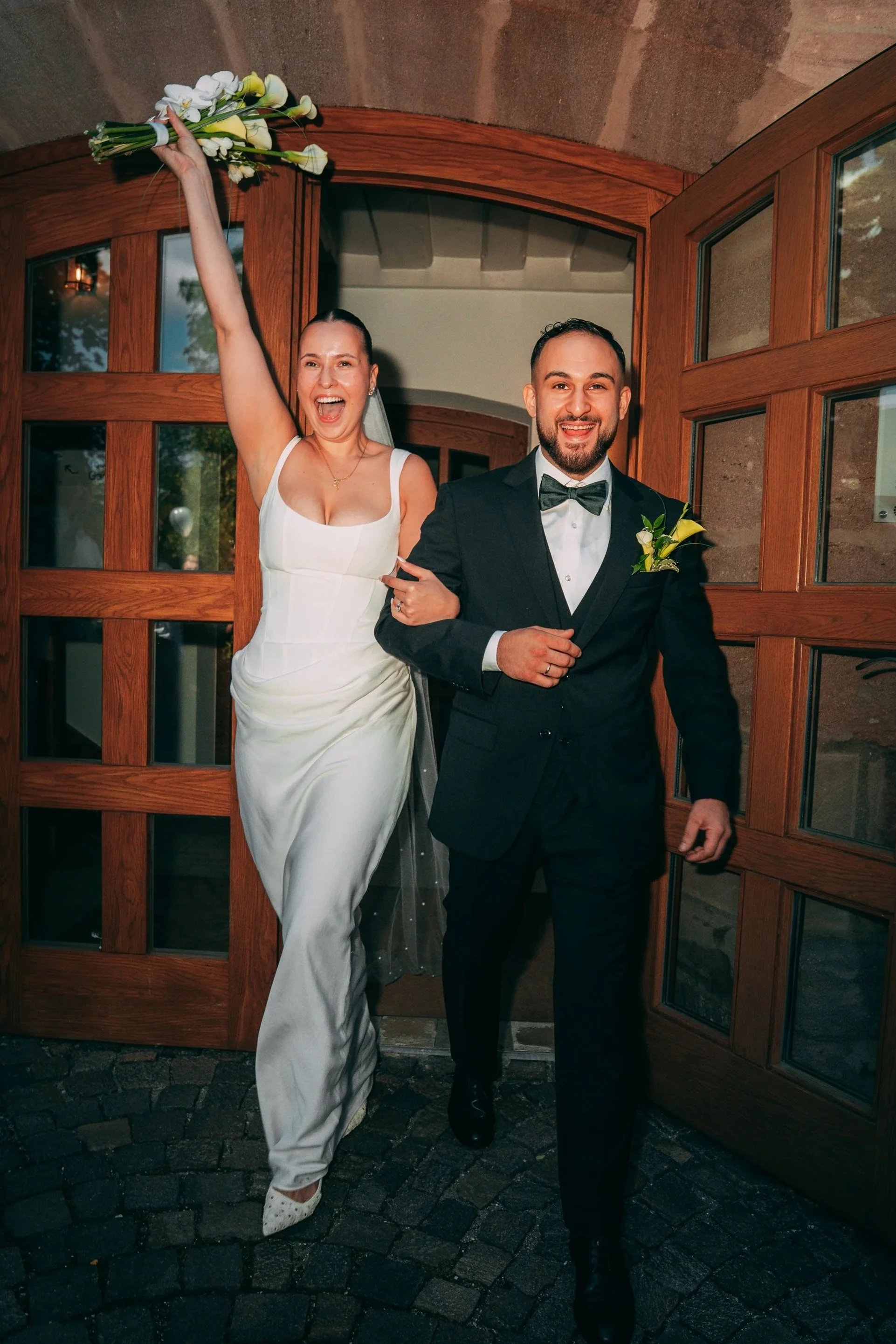 Ein glückliches Hochzeitspaar, die Braut hält einen Blumenstrauß hoch, der Bräutigam trägt einen Anzug mit Fliege und eine Ansteckblume, sie stehen vor einer Holztür auf Kopfsteinpflaster