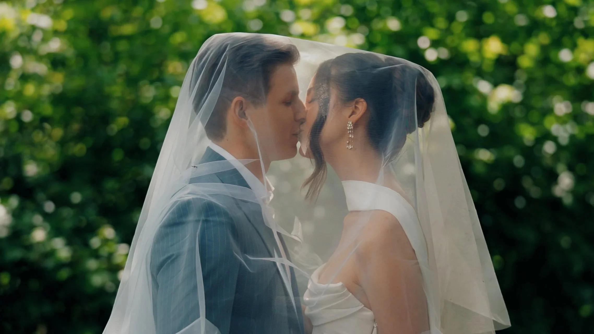 Ein Brautpaar an ihrem Hochzeitstag im Freien, umgeben von grünen Bäumen, die sich küssend unter einem transparenten Schleier.