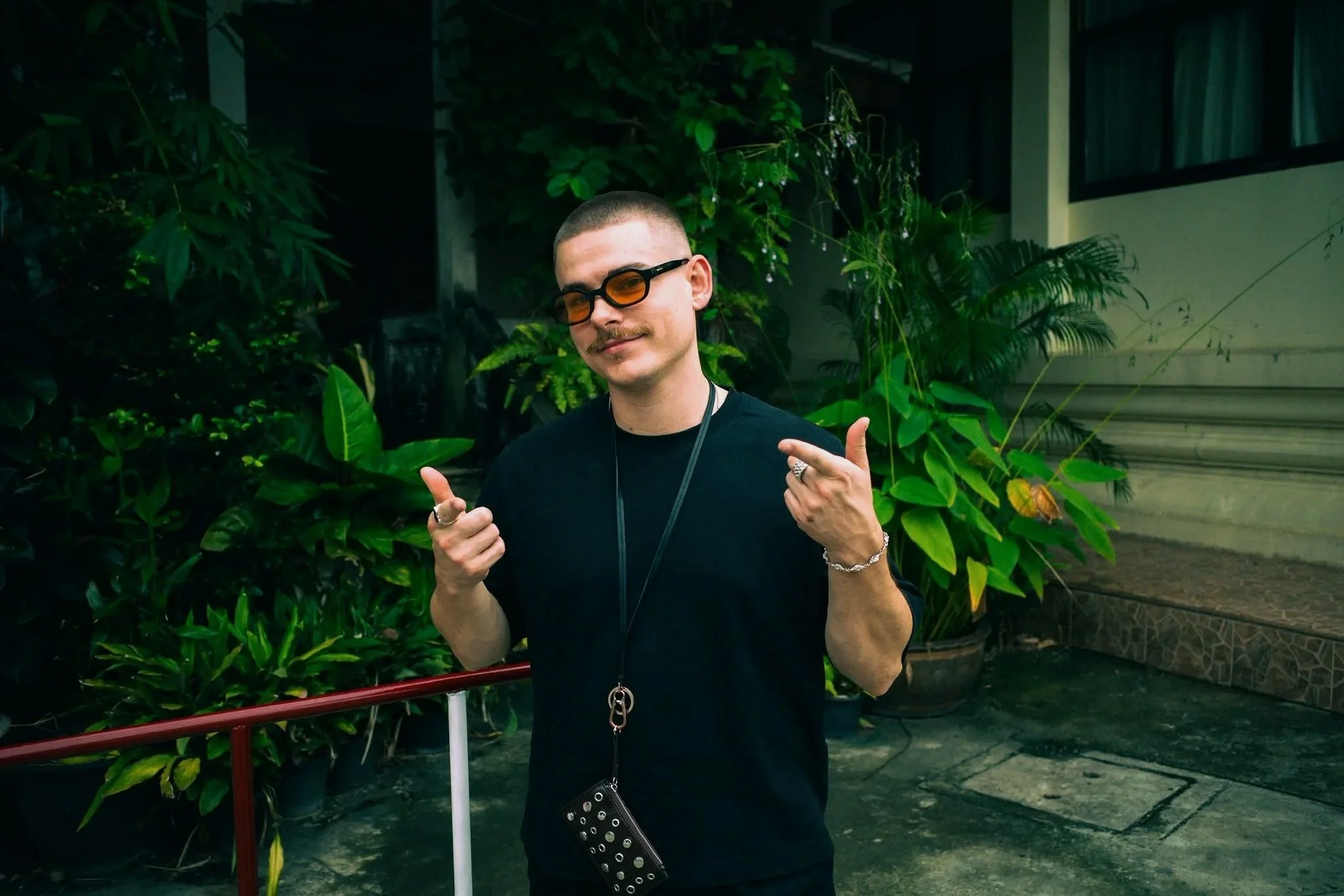 Junge Mann mit kurzen Haaren, Sonnenbrille, schwarzen T-Shirt, Schmuck, posiert im Garten umgeben von grünen Pflanzen.