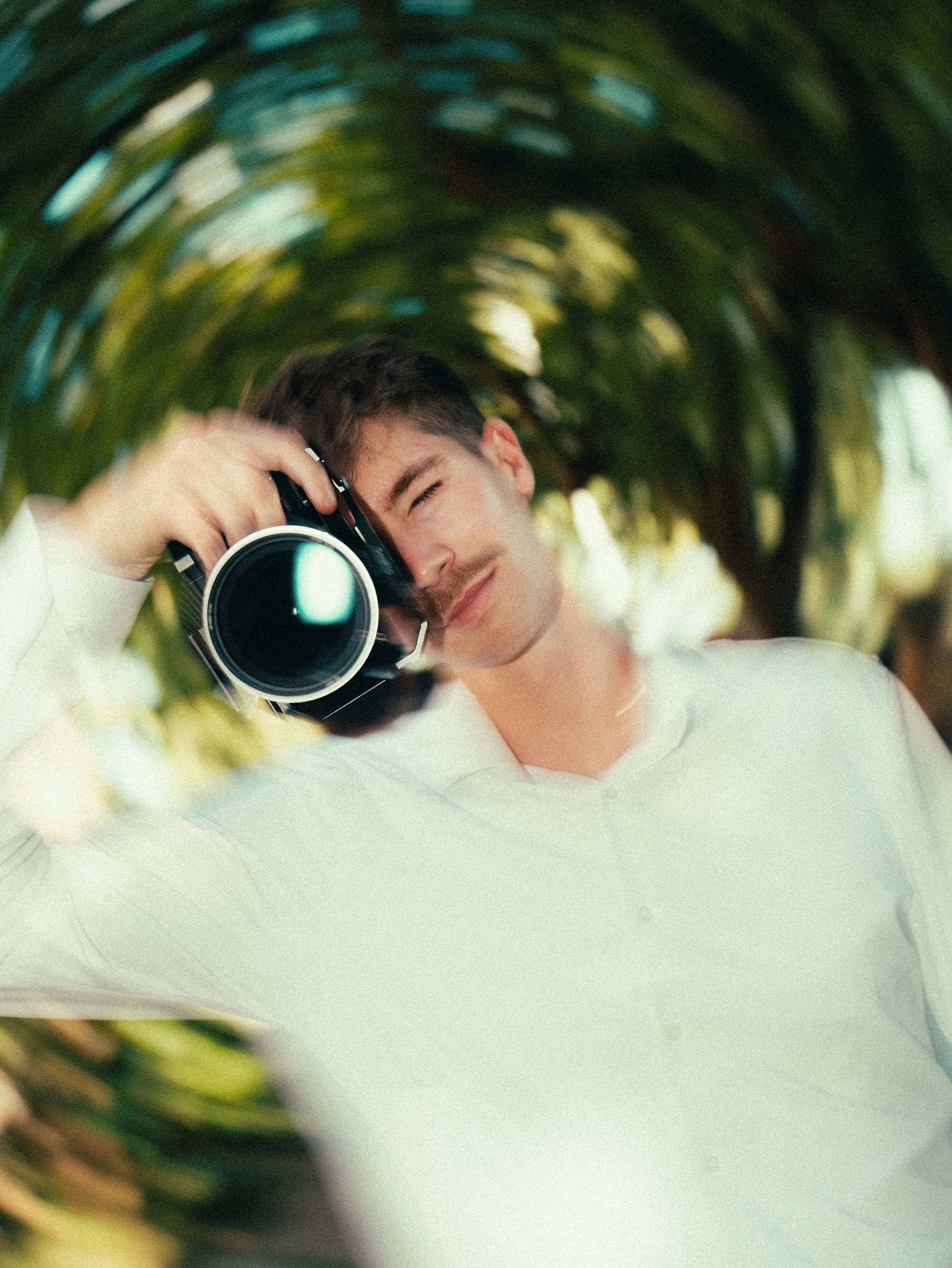 Junger Mann mit dunklem Haar und weißem Hemd fotografiert durch eine grüne Pflanzenröhre, im Hintergrund unscharf. Er hält eine Kamera an sein Gesicht, mit einem breiten Lächeln.