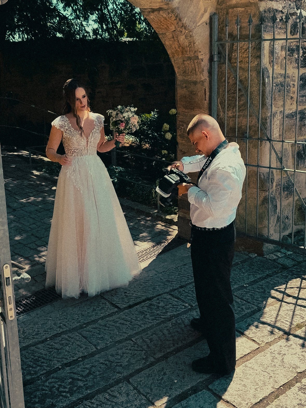 Junge Frau in einem weißen Brautkleid hält einen Blumenstrauß, während ein Mann mit Kamera Fotos macht, vor einem Steinbogen mit Gittertor, im Freien bei Tageslicht.