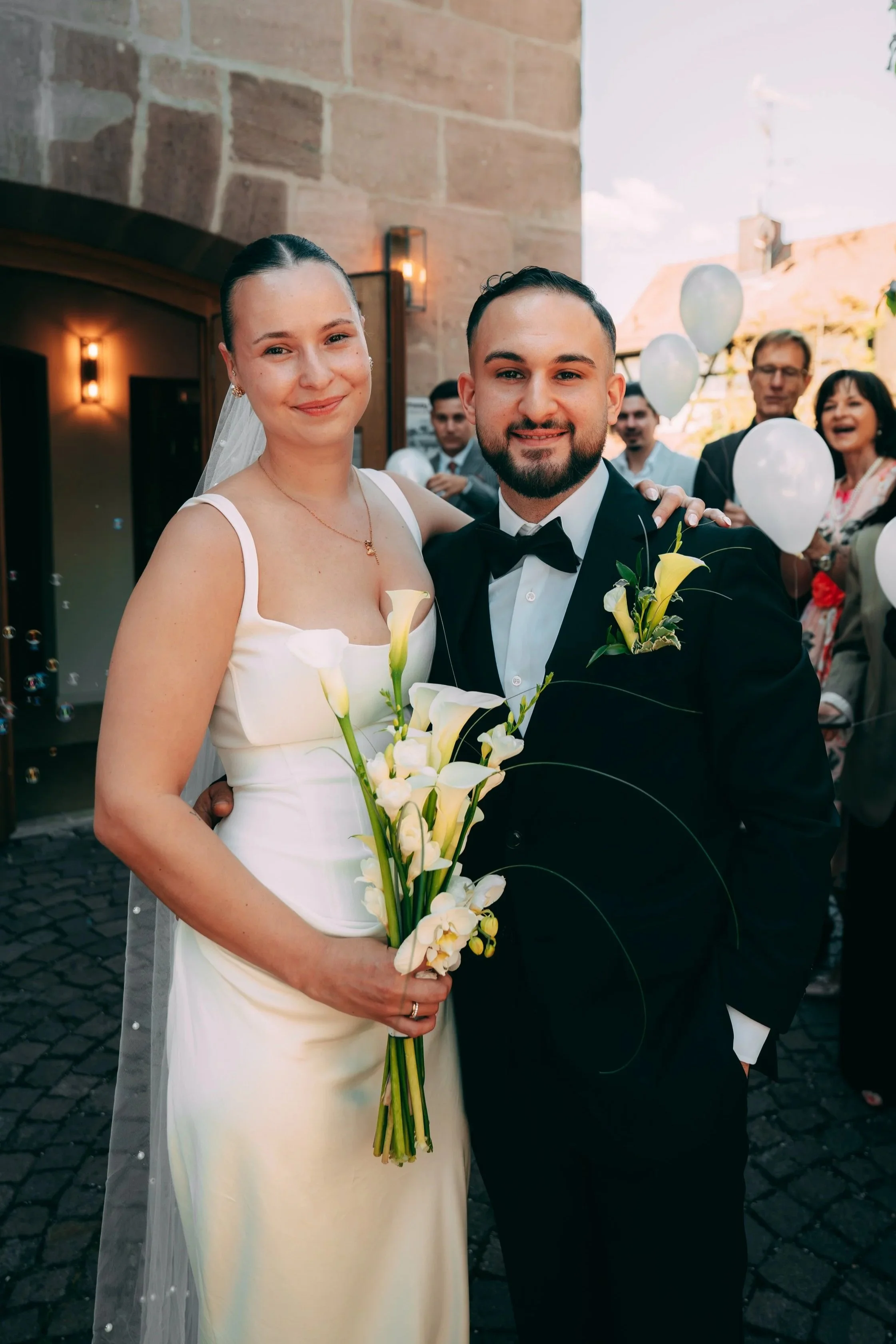 Ein Brautpaar bei einer Hochzeit. Der Bräutigam trägt einen schwarzen Anzug mit Fliege. Hinter ihnen sitzen Gäste, einige halten weiße Ballons, im Hintergrund eine Gebäude mit roten Ziegeldächern.