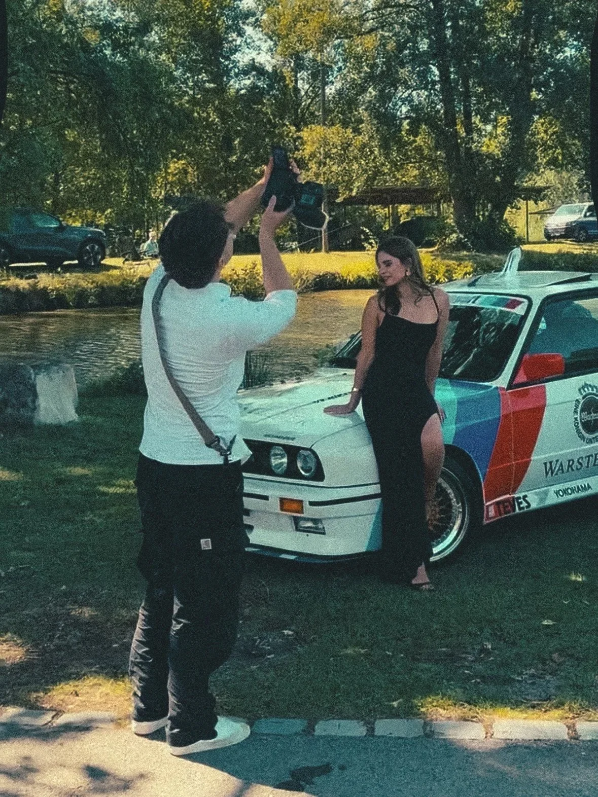 Ein Mann macht ein Foto von einer Frau, die neben einem weißen Auto in einem Park steht. Im Hintergrund sind Bäume und andere Fahrzeuge zu sehen.