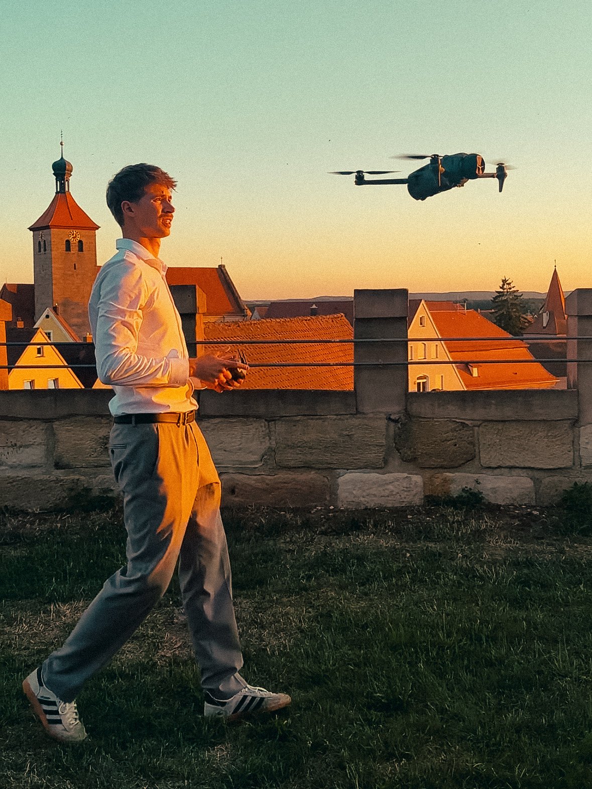 Ein junger Mann steuert eine Drohne mit einer Fernbedienung auf einem Dachgarten bei Sonnenuntergang, im Hintergrund eine Stadt mit Kirchtürmen.