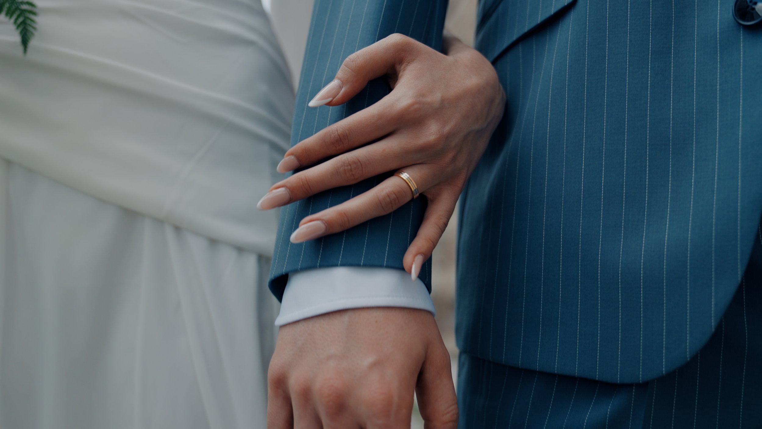 Nahaufnahme einer Hand, die auf den Ärmel einer Person in einem blauen Anzug mit dünnen Streifen zeigt, während eine andere Person in einem weißen Kleid im Hintergrund steht.
