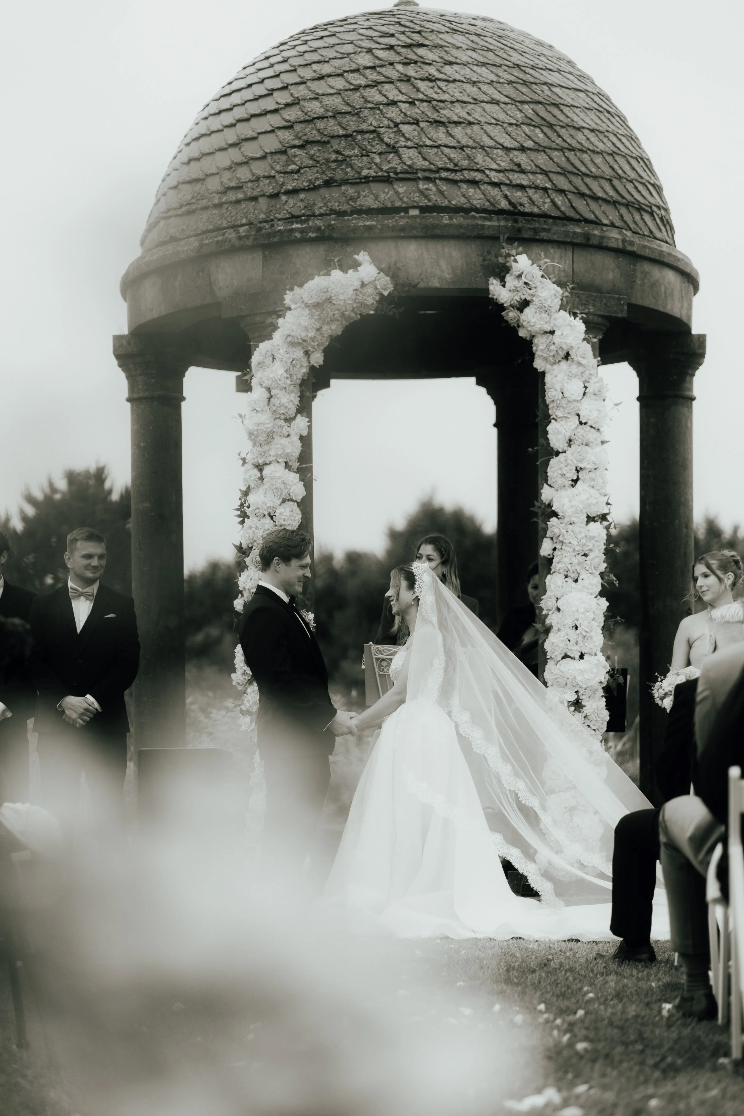 Hochzeitspaar, das sich unter einem Pavillon mit Blumenkranz-Säulen die Eheringe austauscht, umgeben von Gästen, im Freien bei nebligem Wetter.