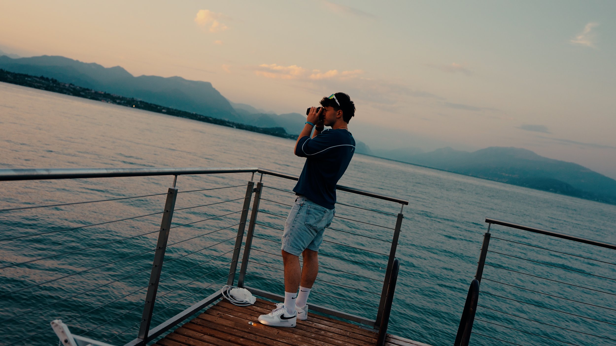 Junge Person mit dunklen Haaren, Sonnenbrille auf dem Kopf, schaut durch ein Fernglas auf einem Boot bei Sonnenuntergang, mit Bergen im Hintergrund.