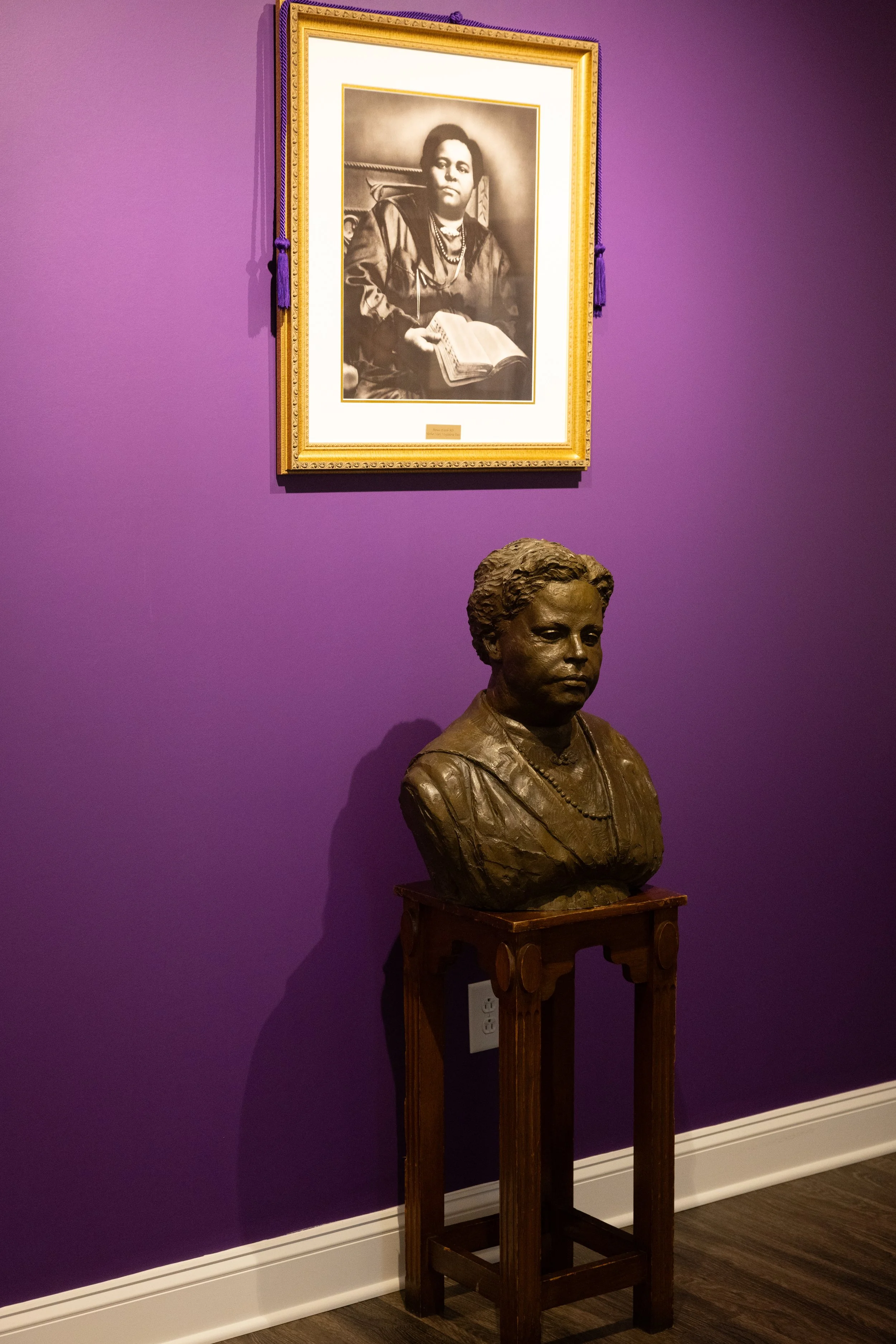 A bronze bust of a woman on a wooden pedestal in a room with purple wall. The wall features a framed black-and-white photograph of a woman holding a book, wearing jewelry, and seated in front of a background with a rope design.