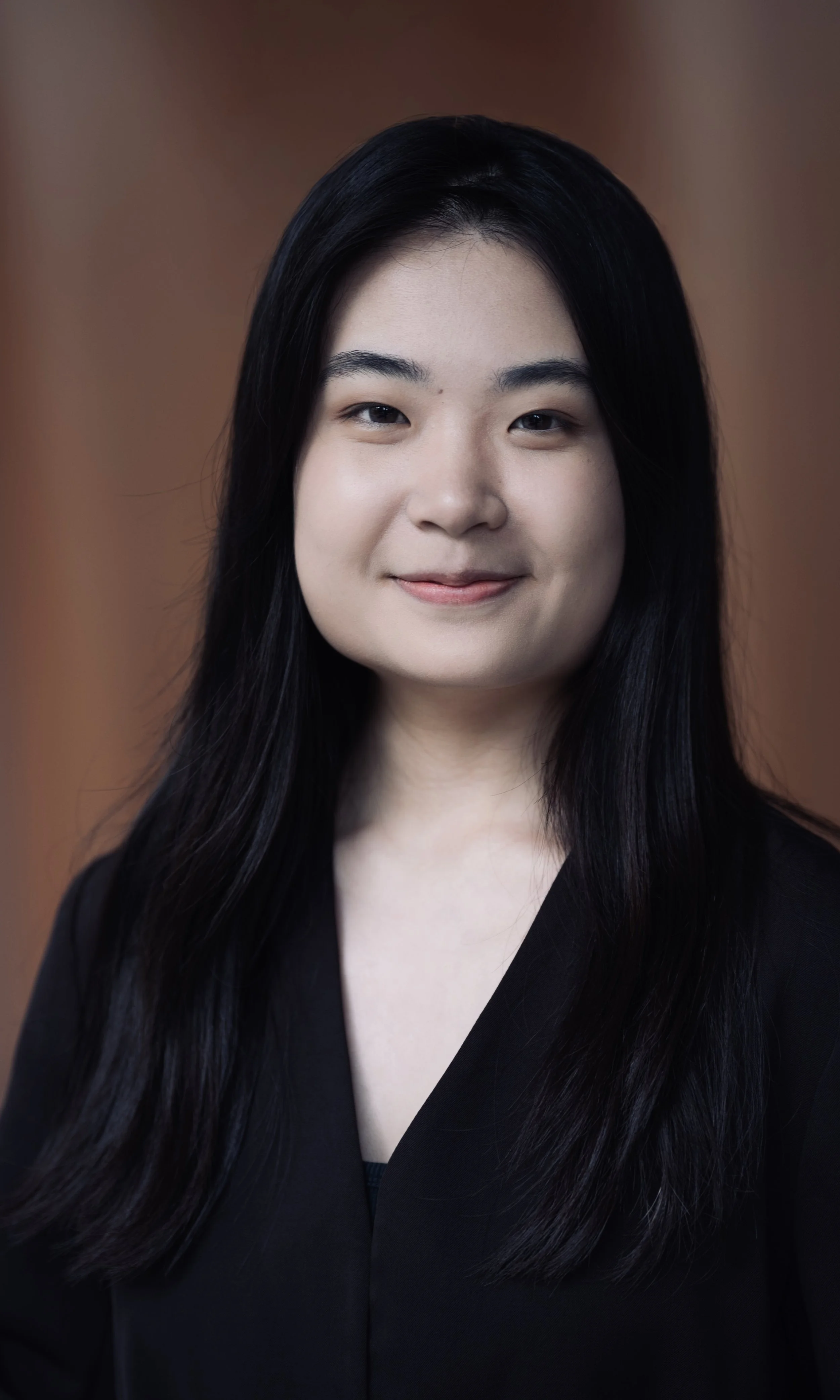 Close-up portrait of an Asian woman with long black hair, smiling softly against a brown background, wearing a black top.