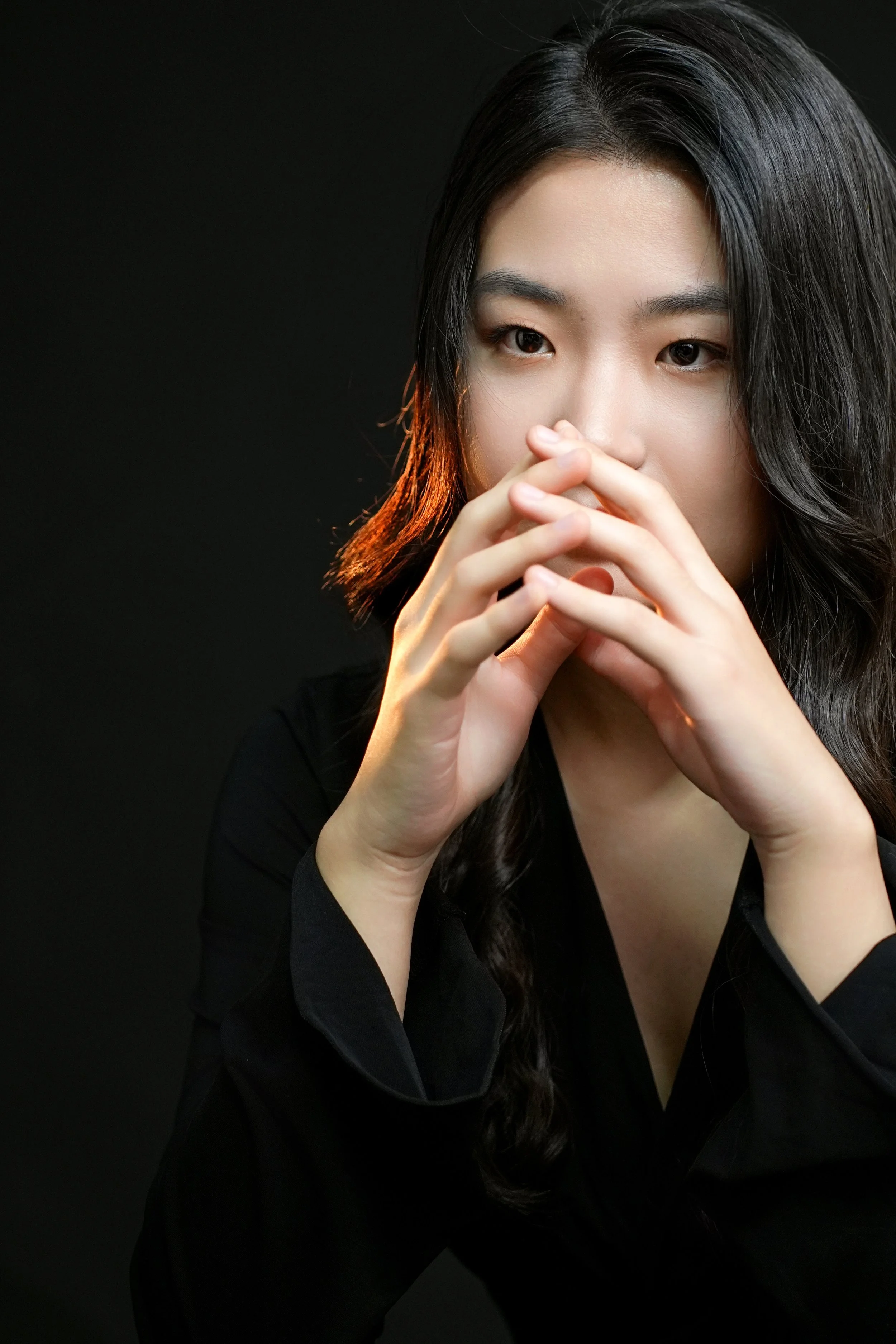 A woman with long dark hair styled in loose waves, wearing a black top and sitting against a black background, with her hands clasped in front of her face.