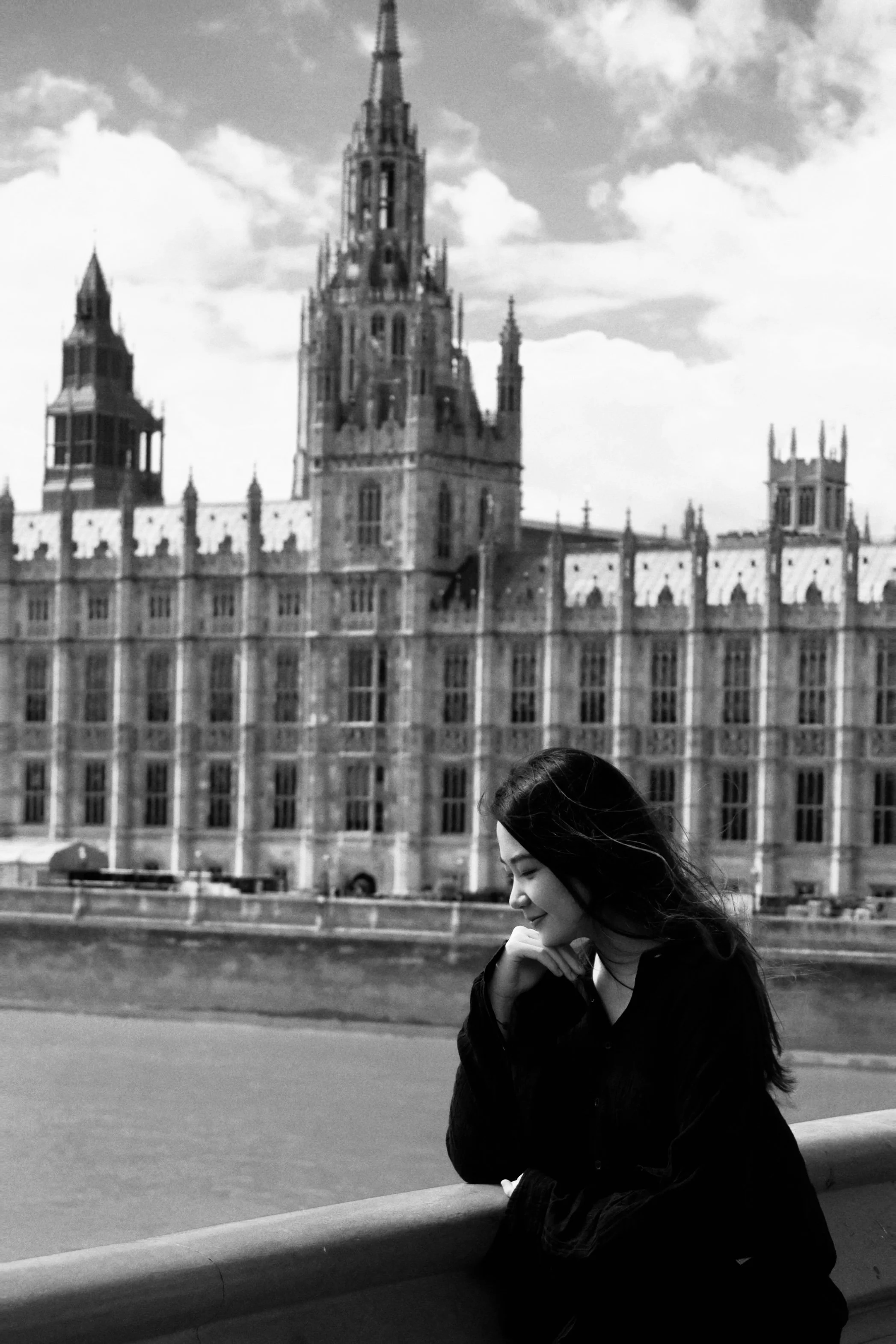 Black and white photo of a young woman with long dark hair, sitting and smiling with her hand near her chin, in front of the Palace of Westminster and Big Ben in London, UK.